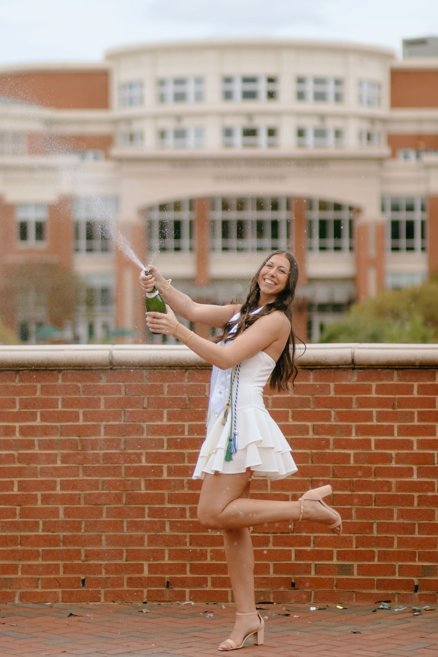 A person in a white dress strikes celebratory poses on a brick wall.
