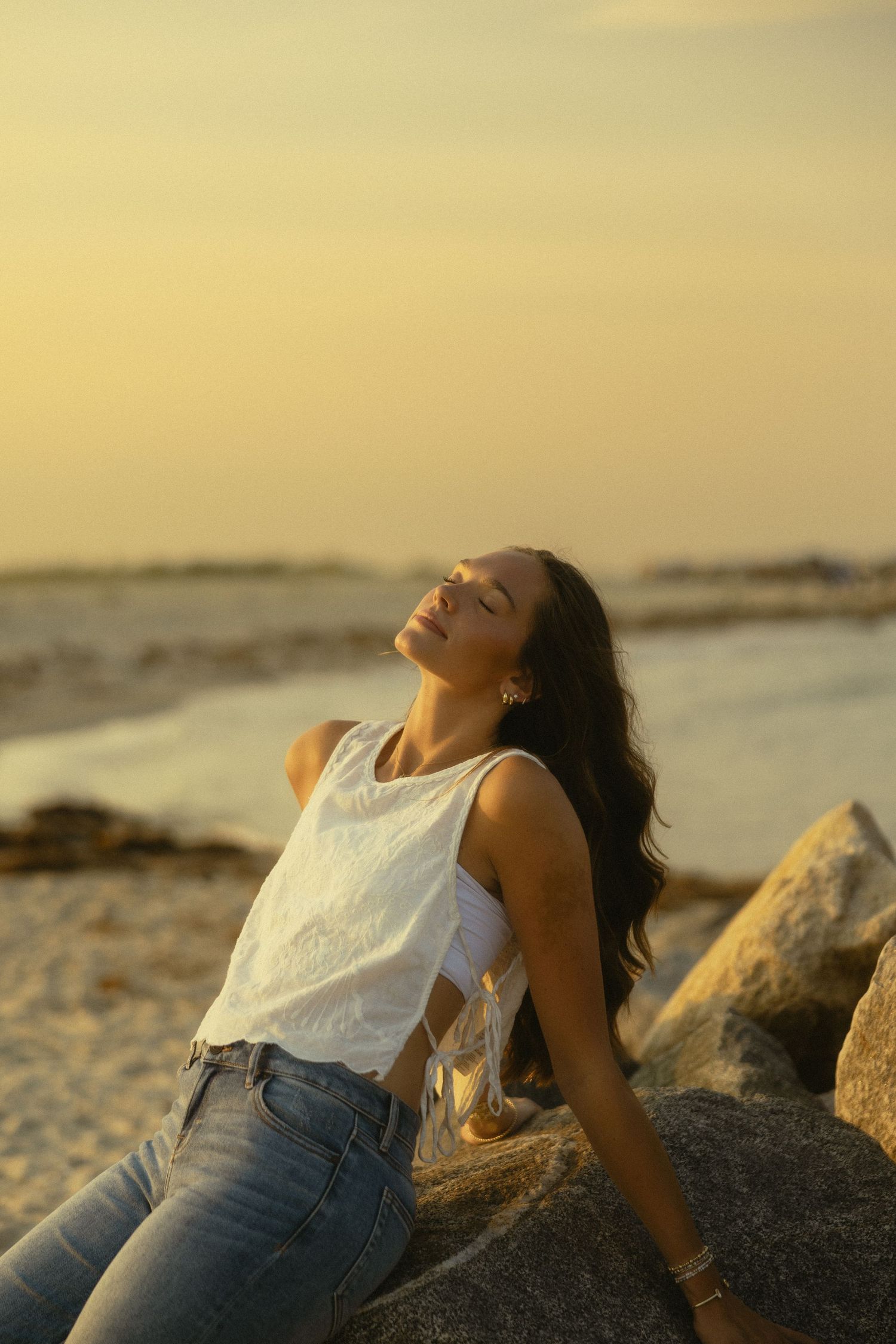 Young woman in white top and jeans relaxing on beach rocks at golden sunset with eyes closed.