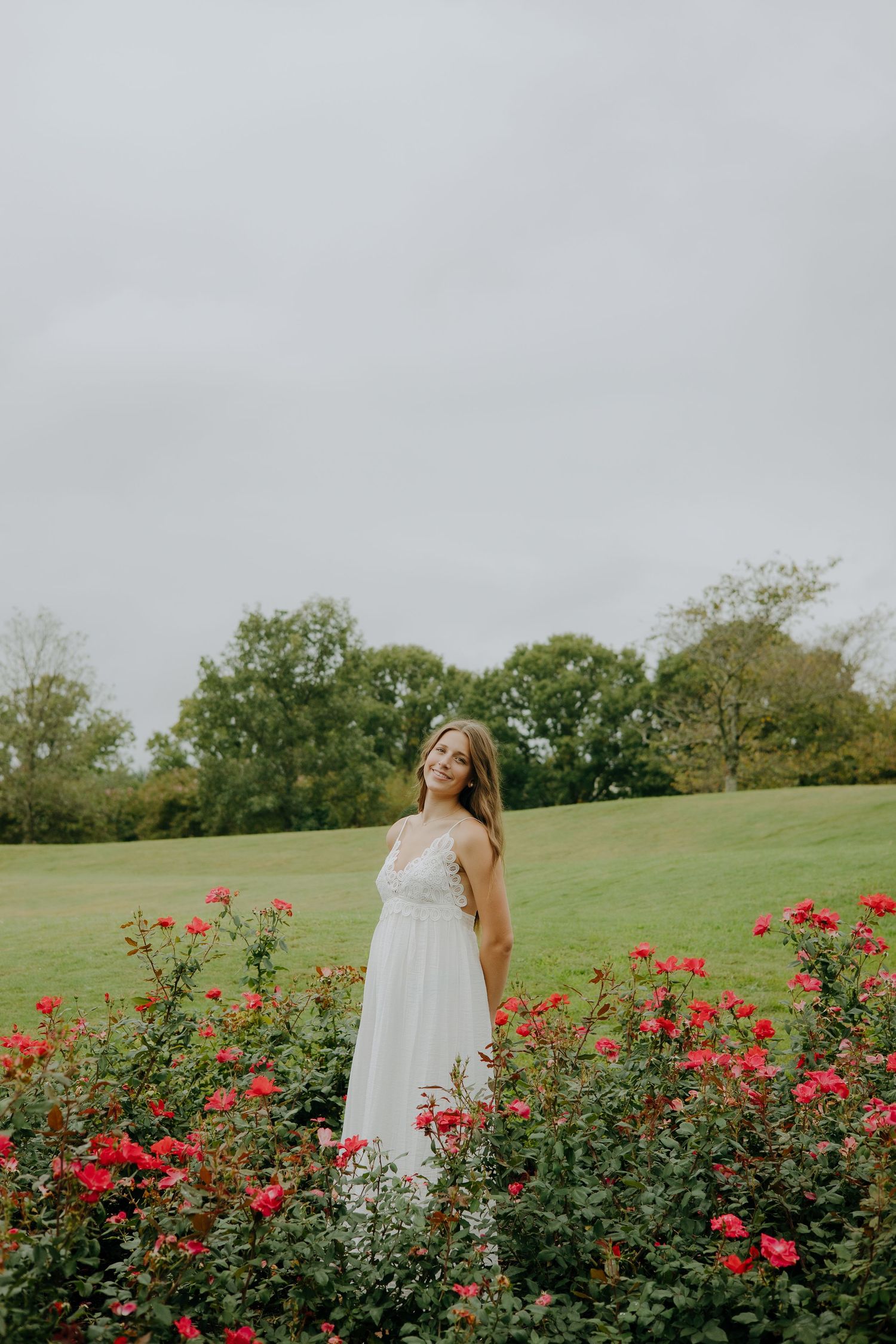 A young woman in a white lace dress stands among red roses in a lush green garden on an overcast day.