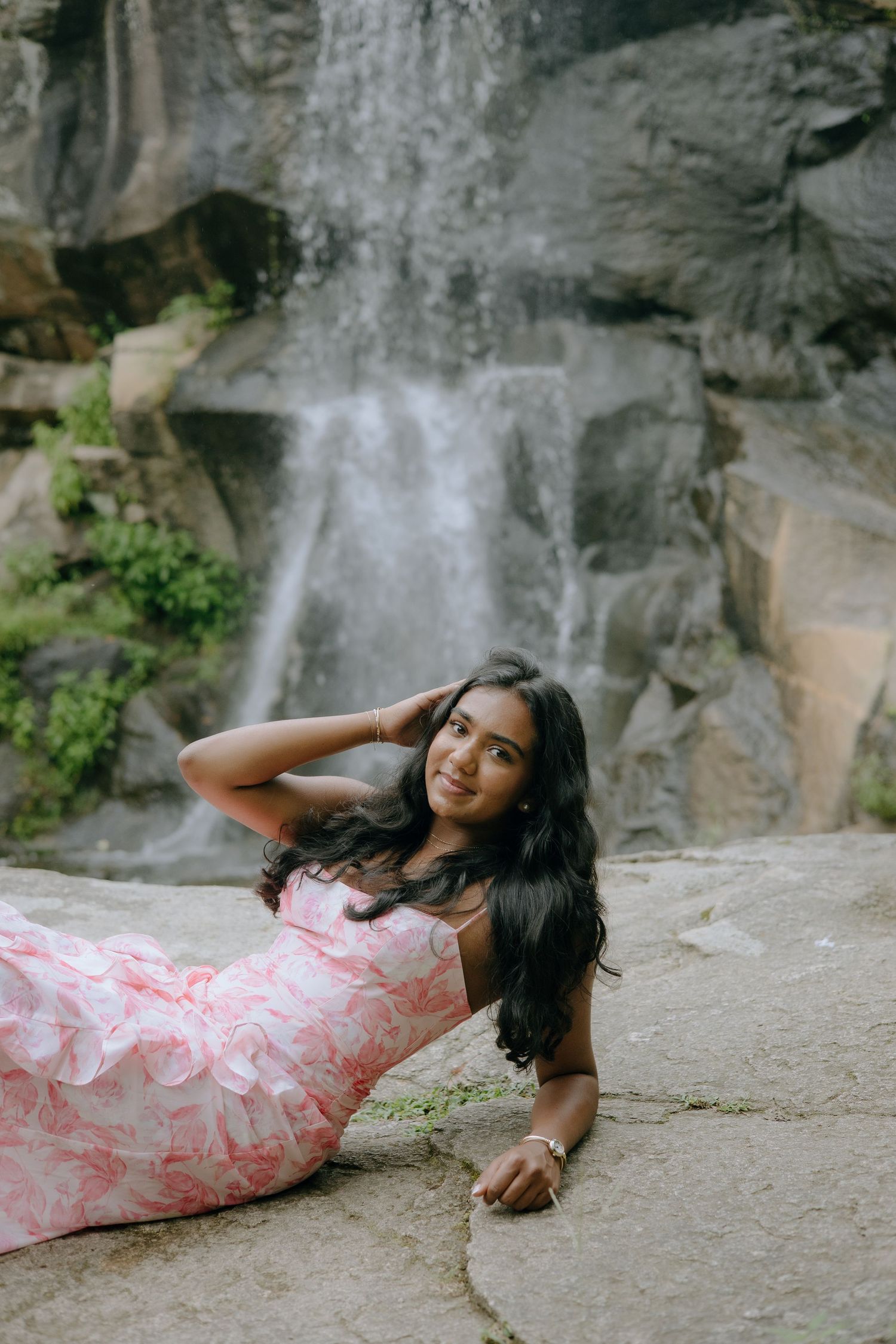 Young woman in pink floral dress posing gracefully on rocks in front of a cascading waterfall.