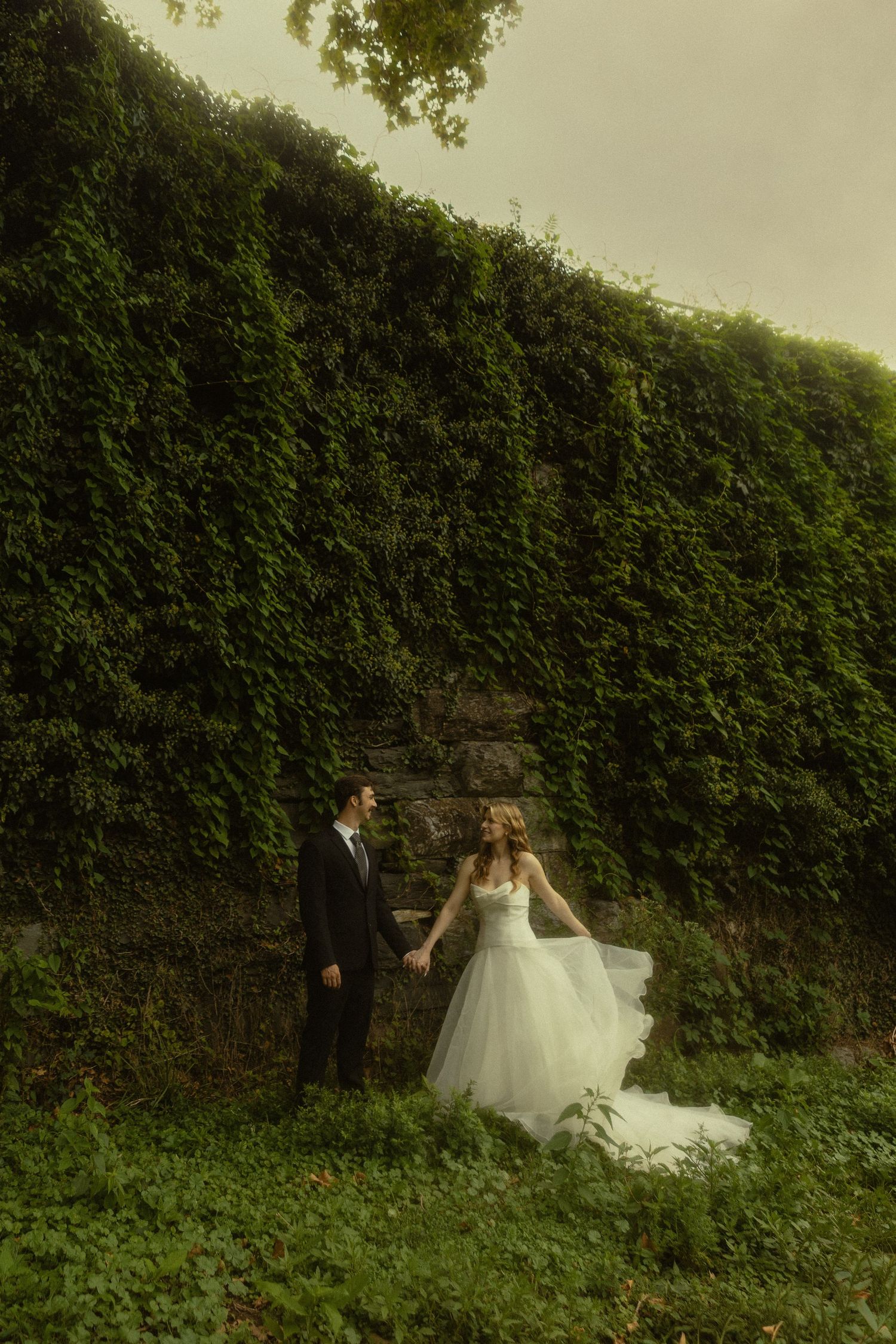 Bride and groom hold hands before an ivy-covered stone wall, bride in flowing white gown, groom in black suit.