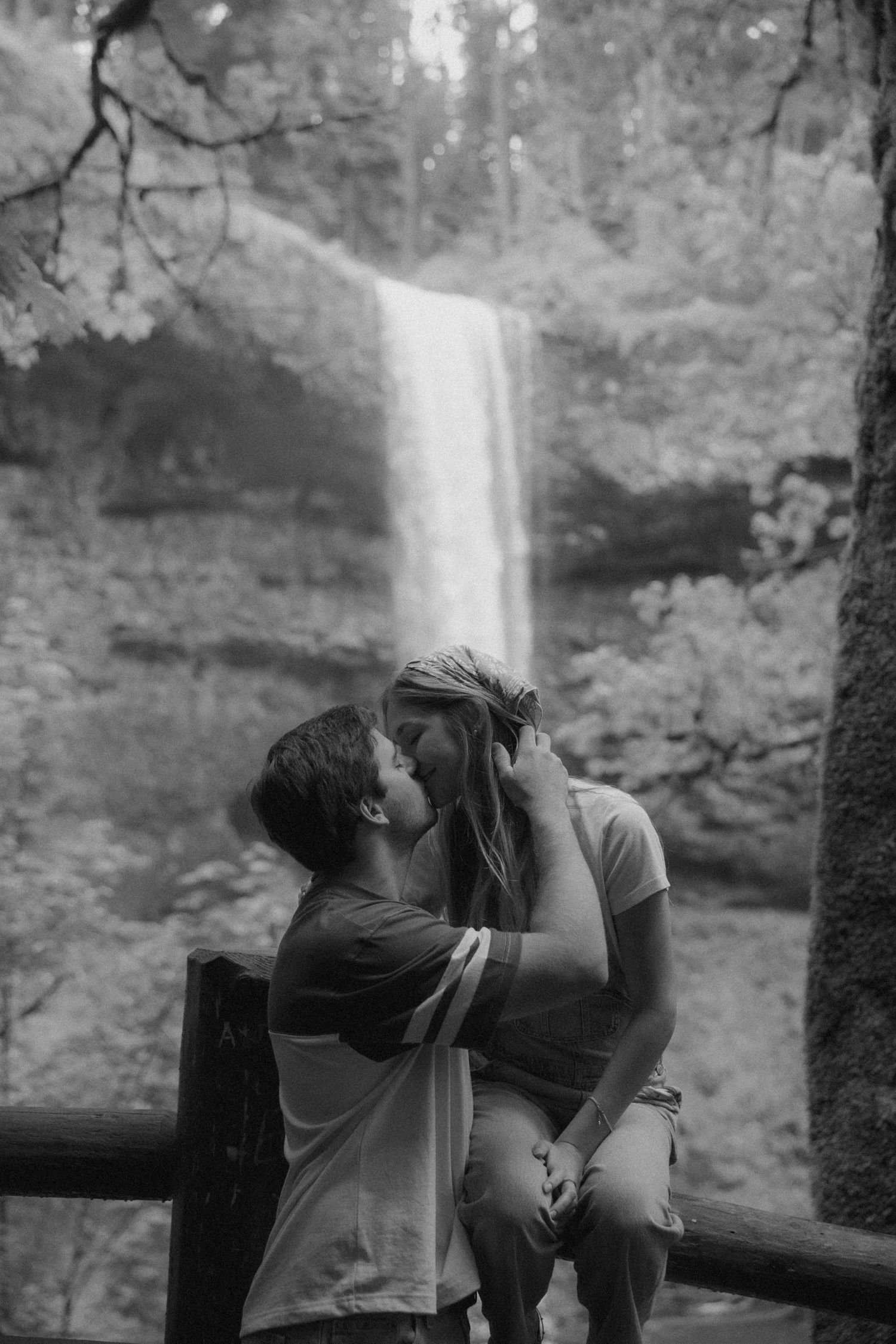 Black and white photo of a couple sharing a kiss on a fence with a stunning waterfall and forest backdrop.