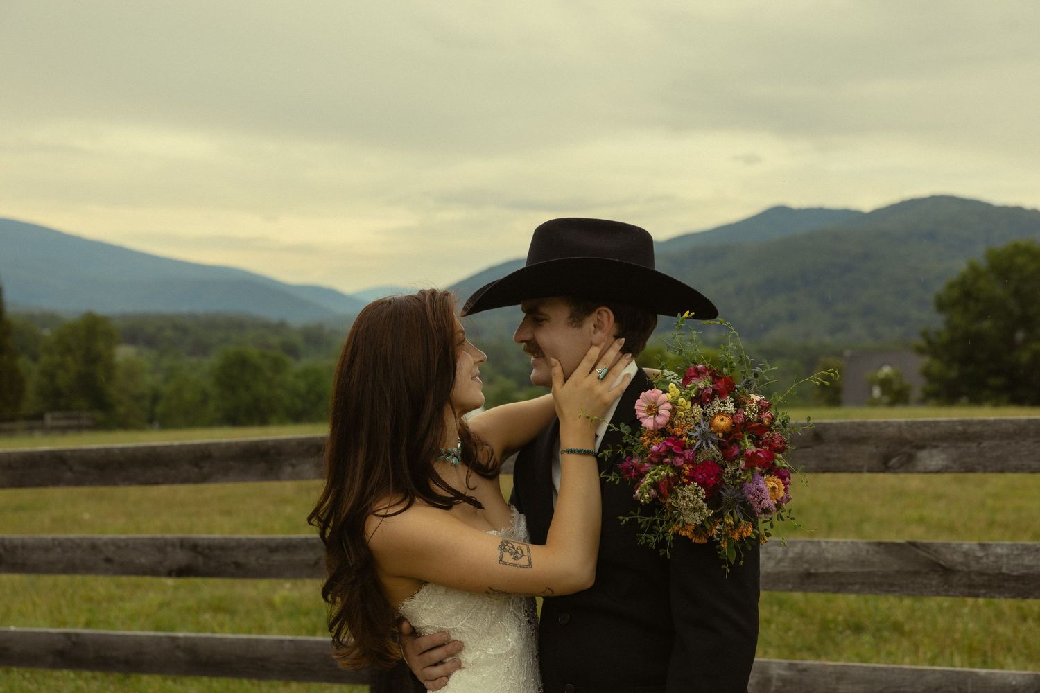 Bride and groom sharing a romantic moment outdoors, groom in black cowboy hat holding colorful bouquet, mountains in background.