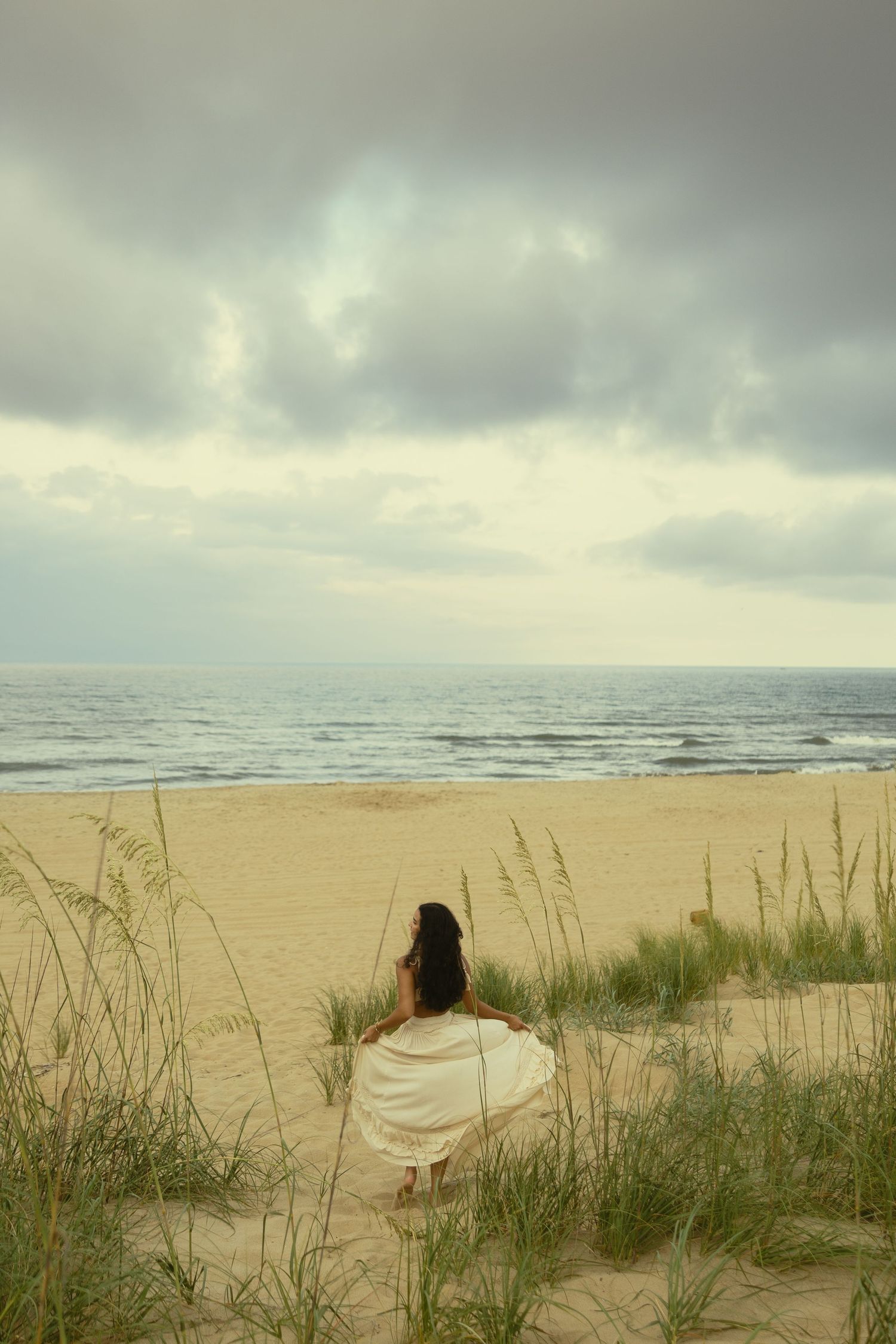 Woman in white dress sitting on sandy beach dunes overlooking calm ocean under cloudy sky.