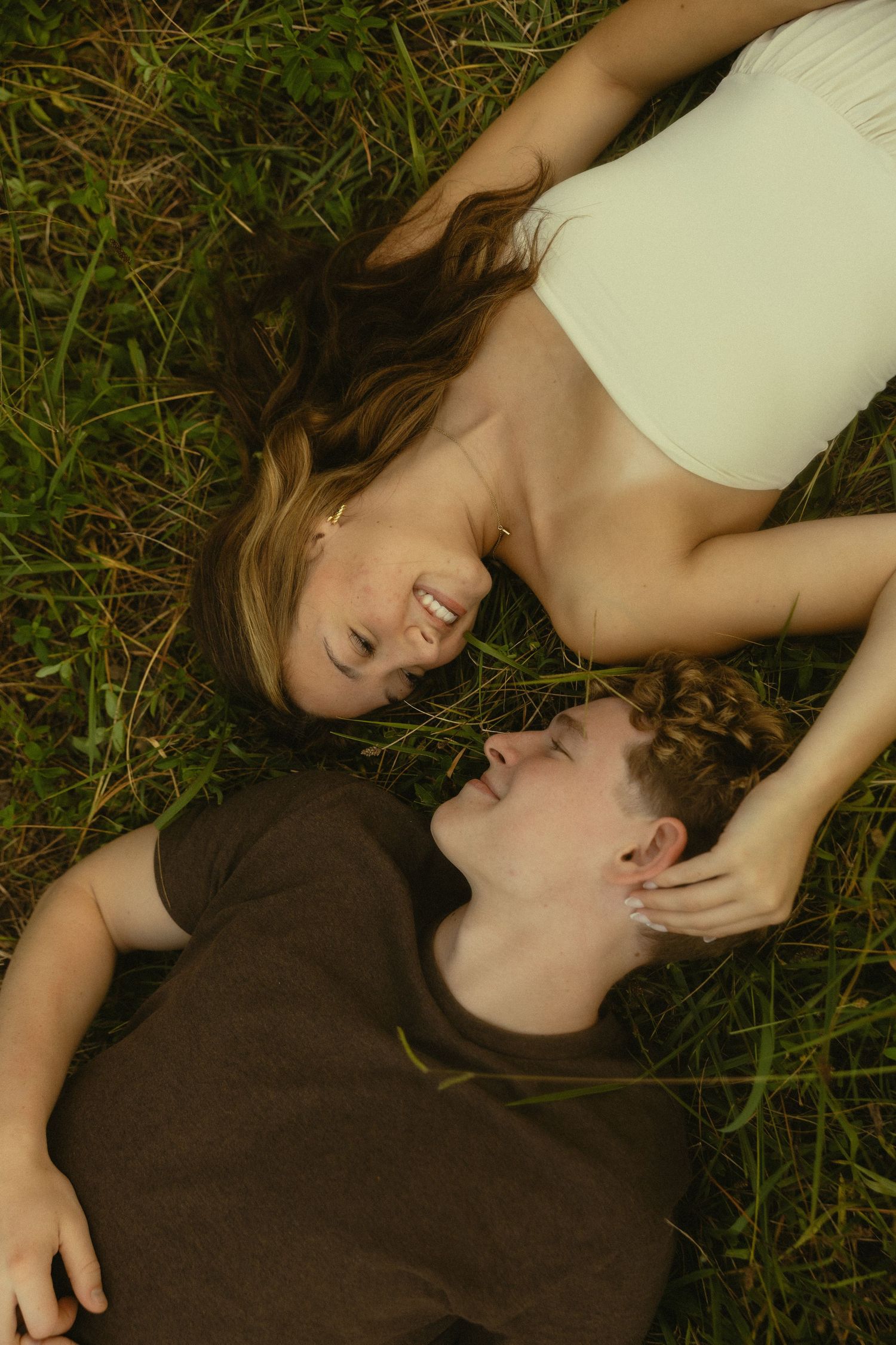 Young couple lying in green grass, smiling and gazing at each other on a sunny day outdoors.