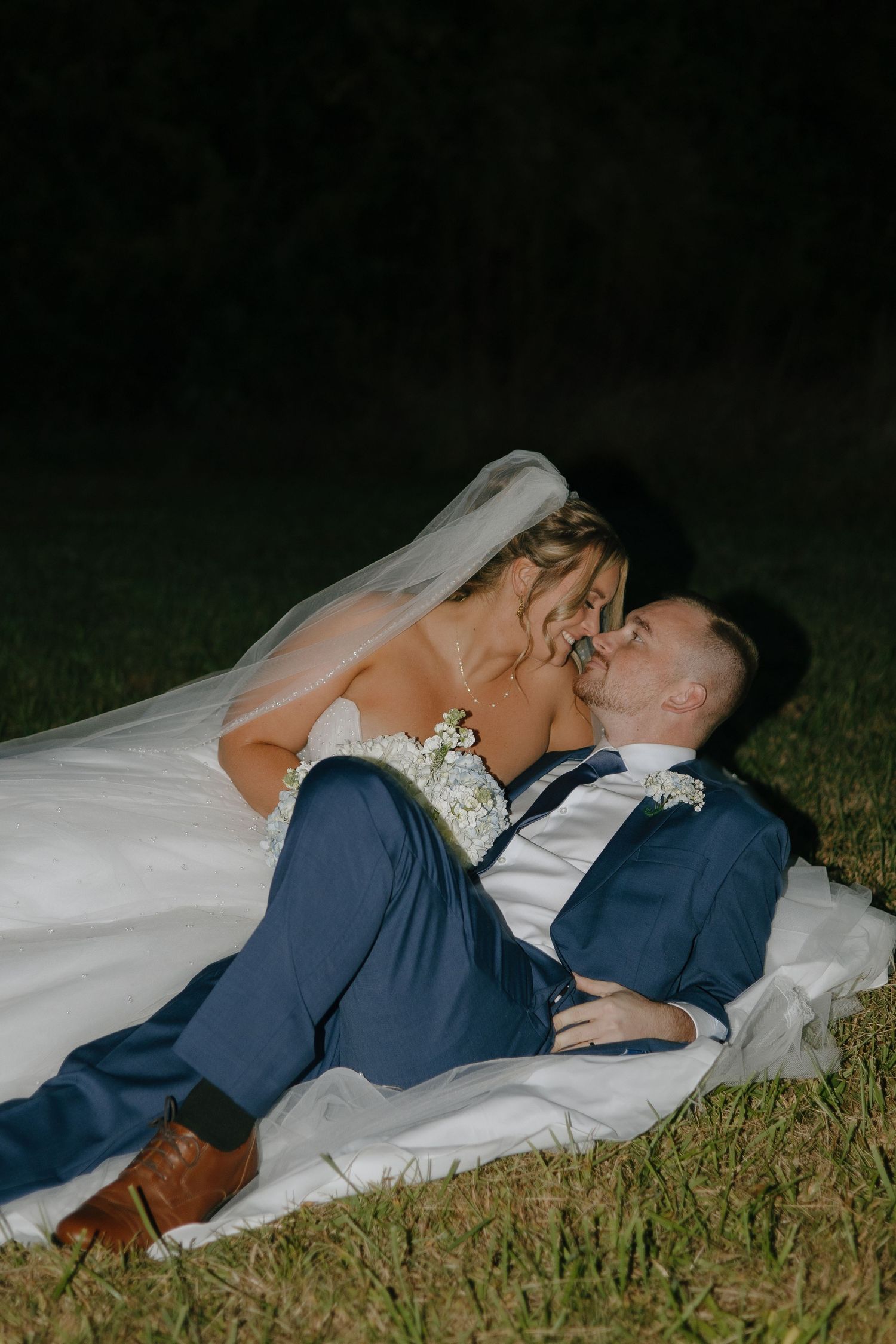 A romantic wedding couple lies on a blanket in the grass at dusk, with the bride's veil flowing behind her.