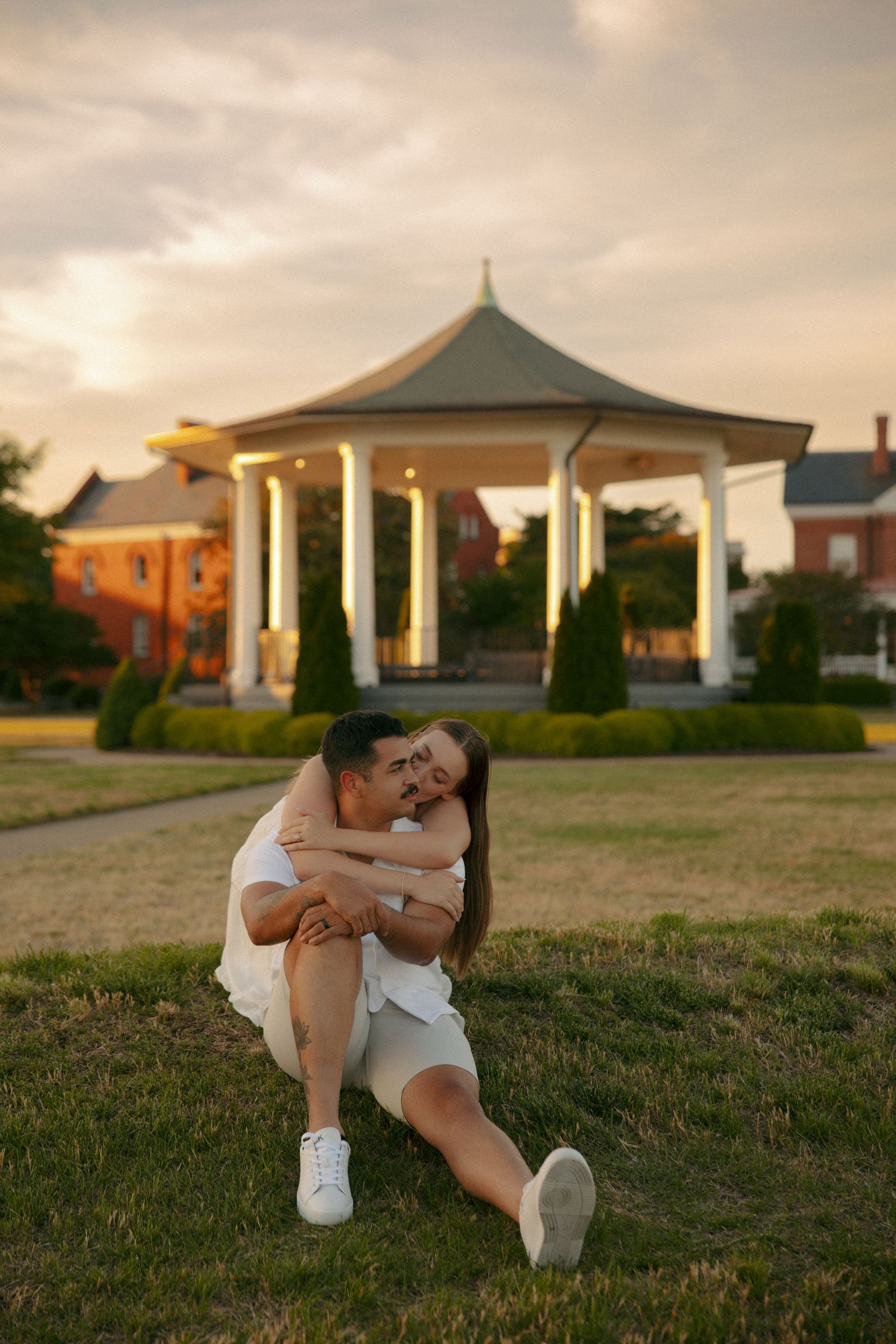 Couple embracing on grass at sunset near a white gazebo, man lifting woman in a romantic outdoor park setting.