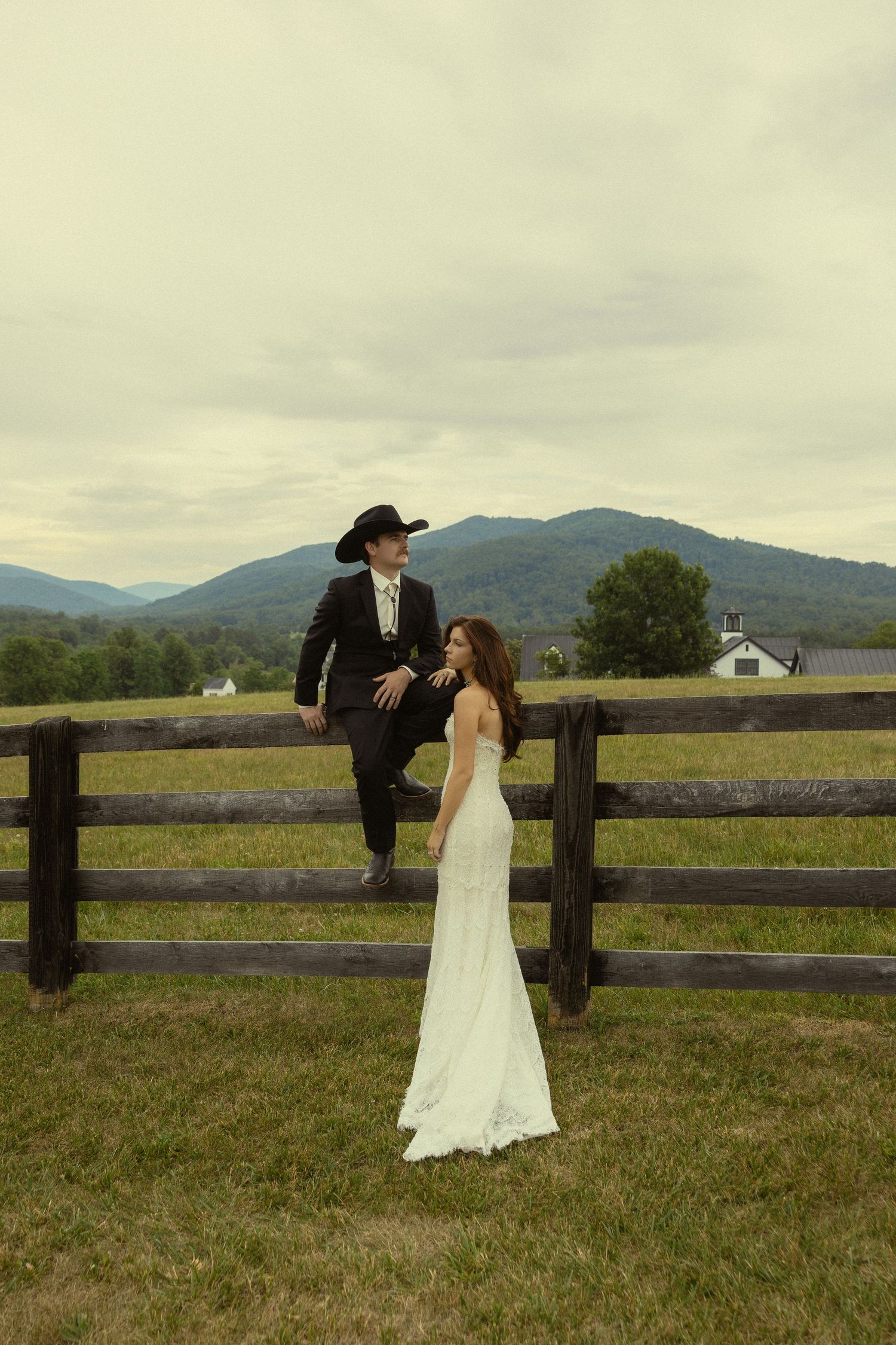 Bride in white gown and groom in black suit with cowboy hat pose by rustic fence with mountain backdrop.