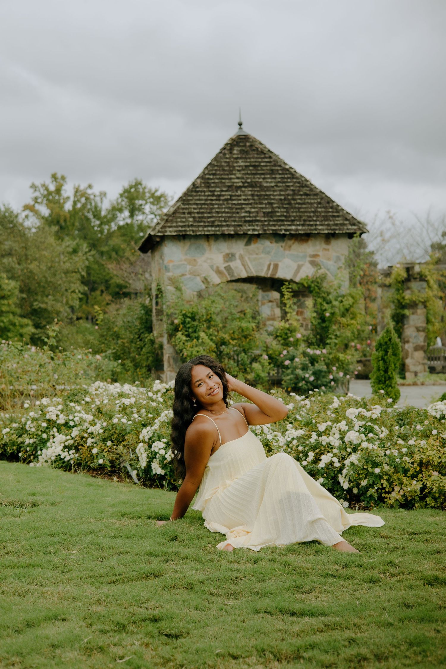 A person in a flowing cream dress reclines gracefully on lush green grass near blooming flowers in a garden setting.