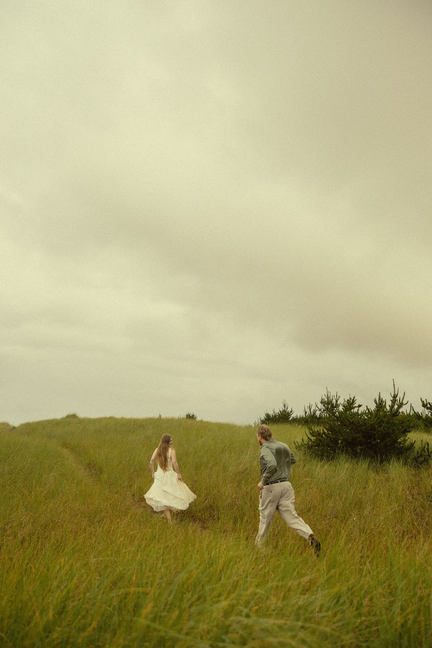 A bride in a white dress and groom in gray suit run through a lush green meadow under an overcast sky.