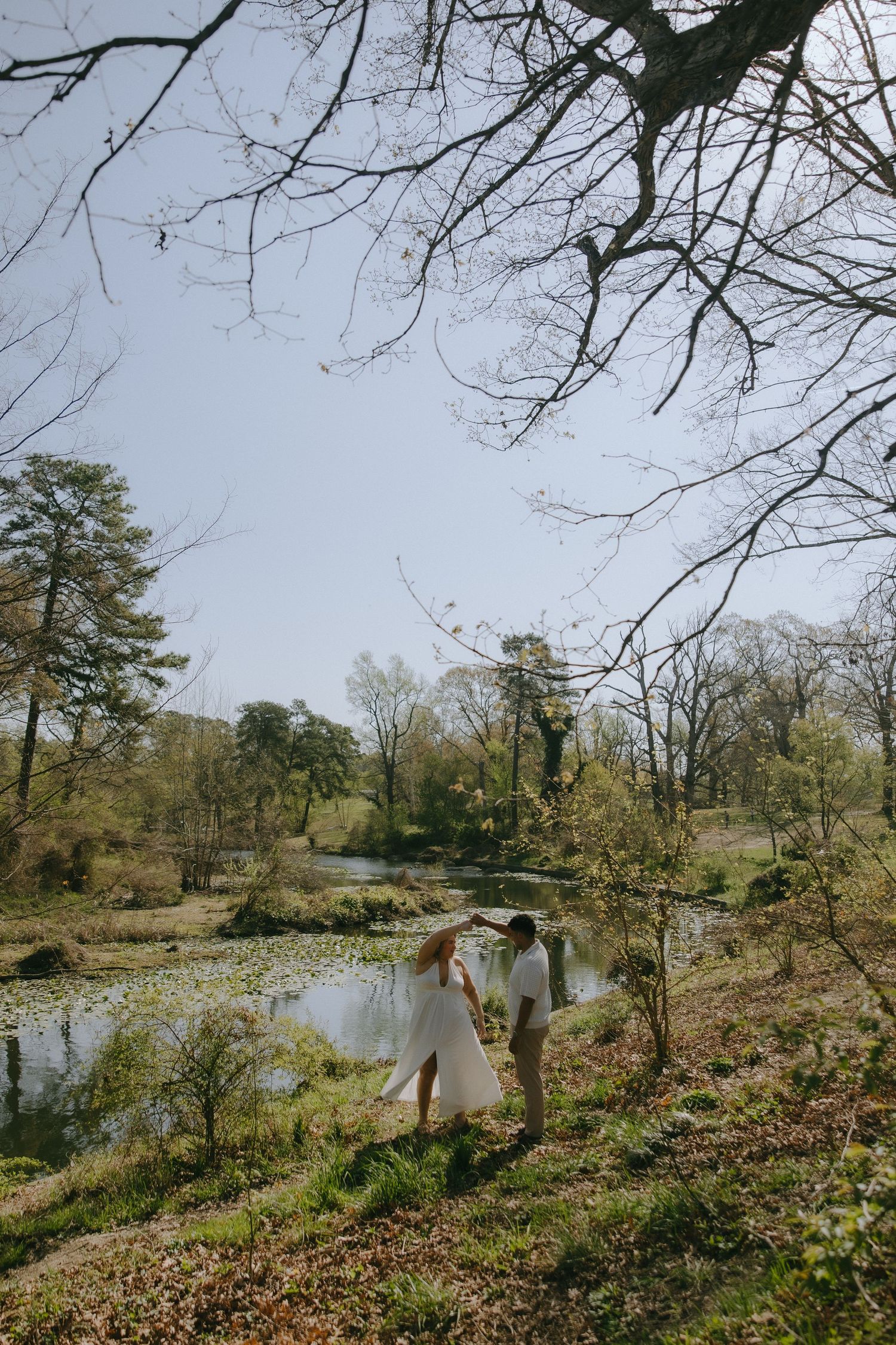 Bride and groom hold hands near a serene pond surrounded by lush trees in an outdoor wedding setting.