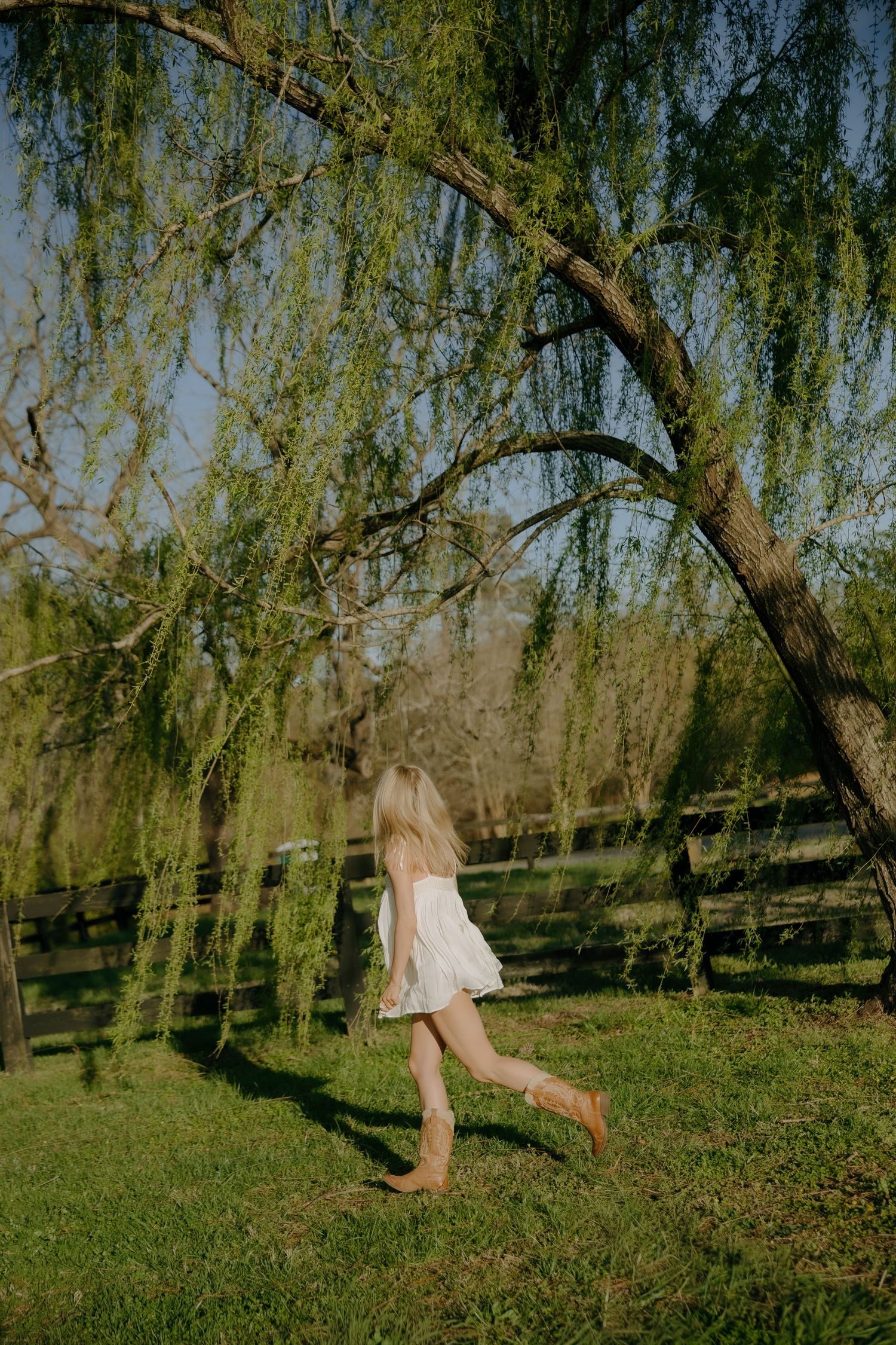 A barefoot girl in a white dress runs through lush green grass beneath a weeping willow tree.