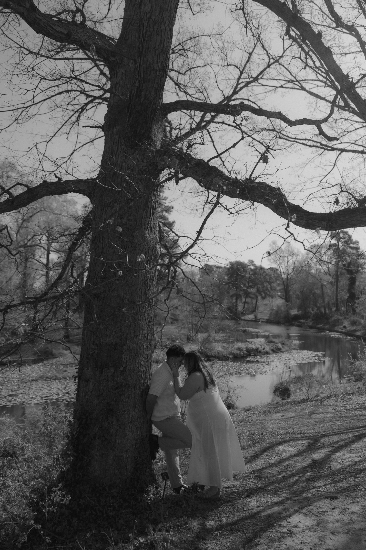 Black and white photo of a couple in white attire sharing a romantic moment under a large bare tree by a serene pond.