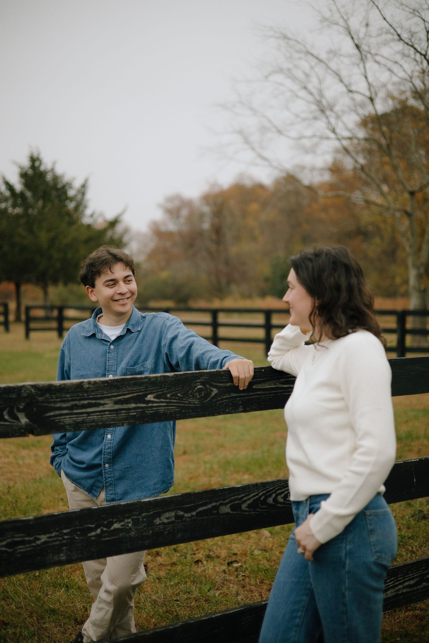 A couple sharing a casual moment while leaning on a wooden fence in an autumn field.
