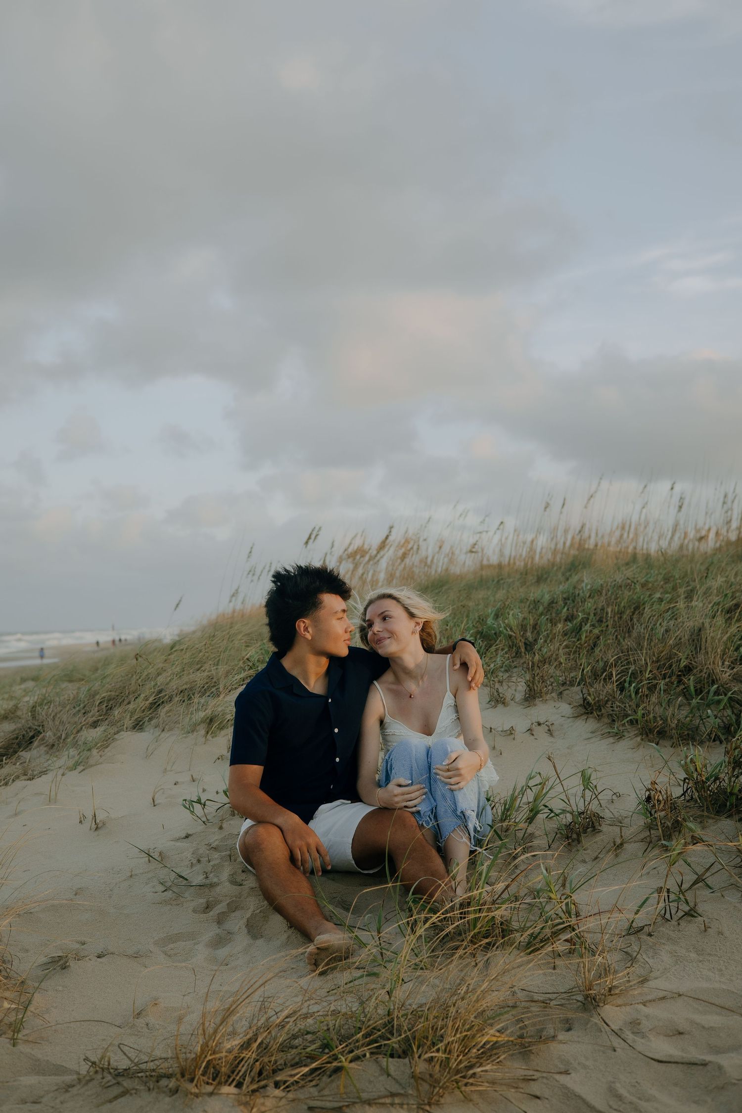 A romantic couple embraces on sandy beach dunes at sunset with tall grass swaying in the breeze.