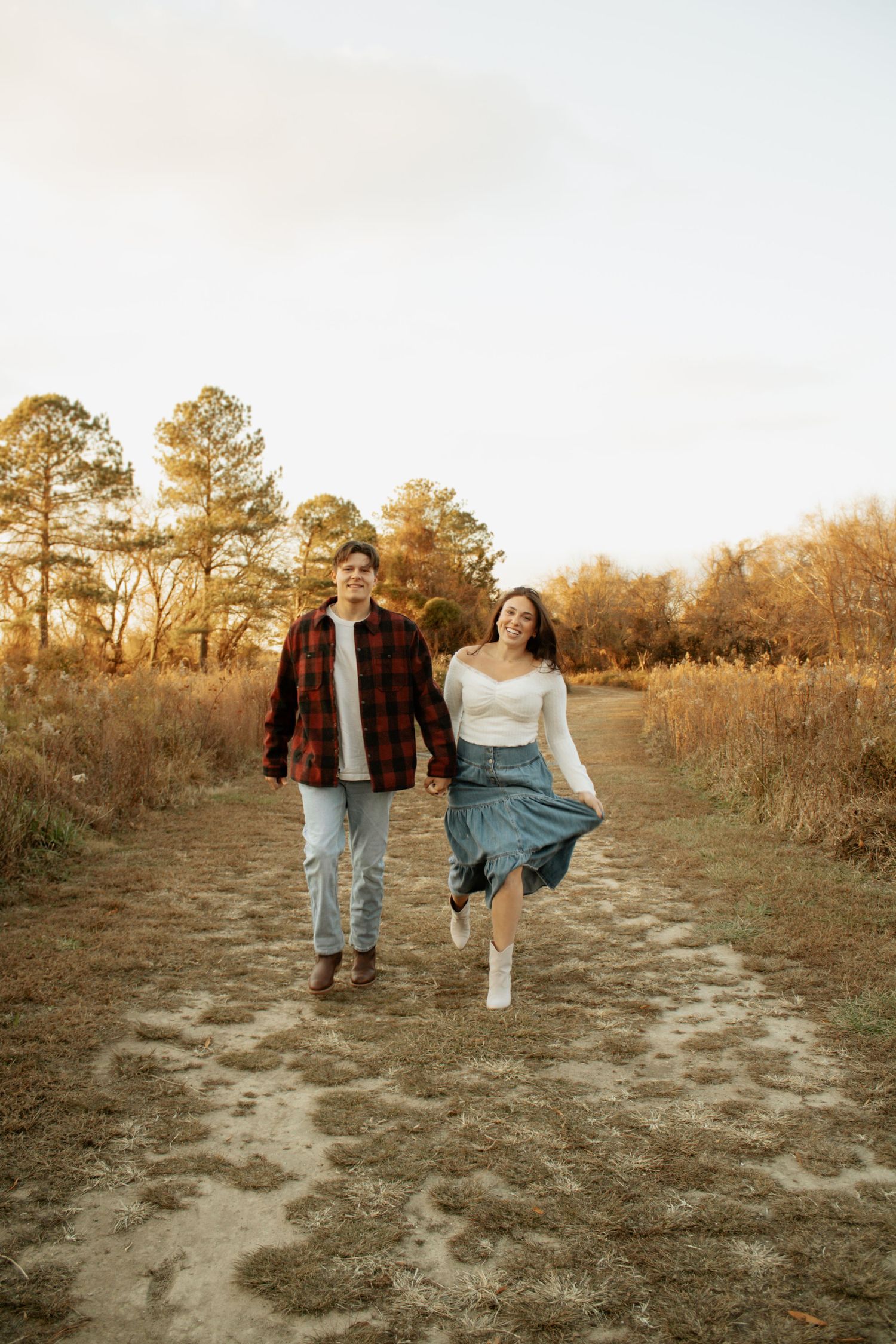 A couple walks along a dirt path during golden hour with pine trees in the background.