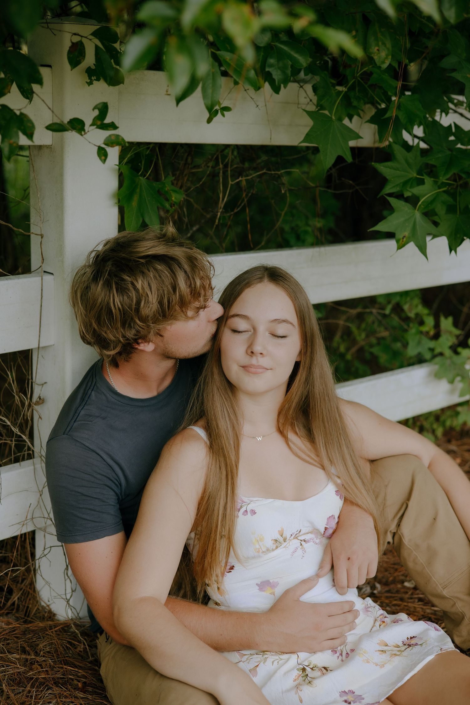 A couple shares an intimate moment while sitting against a white fence surrounded by greenery.