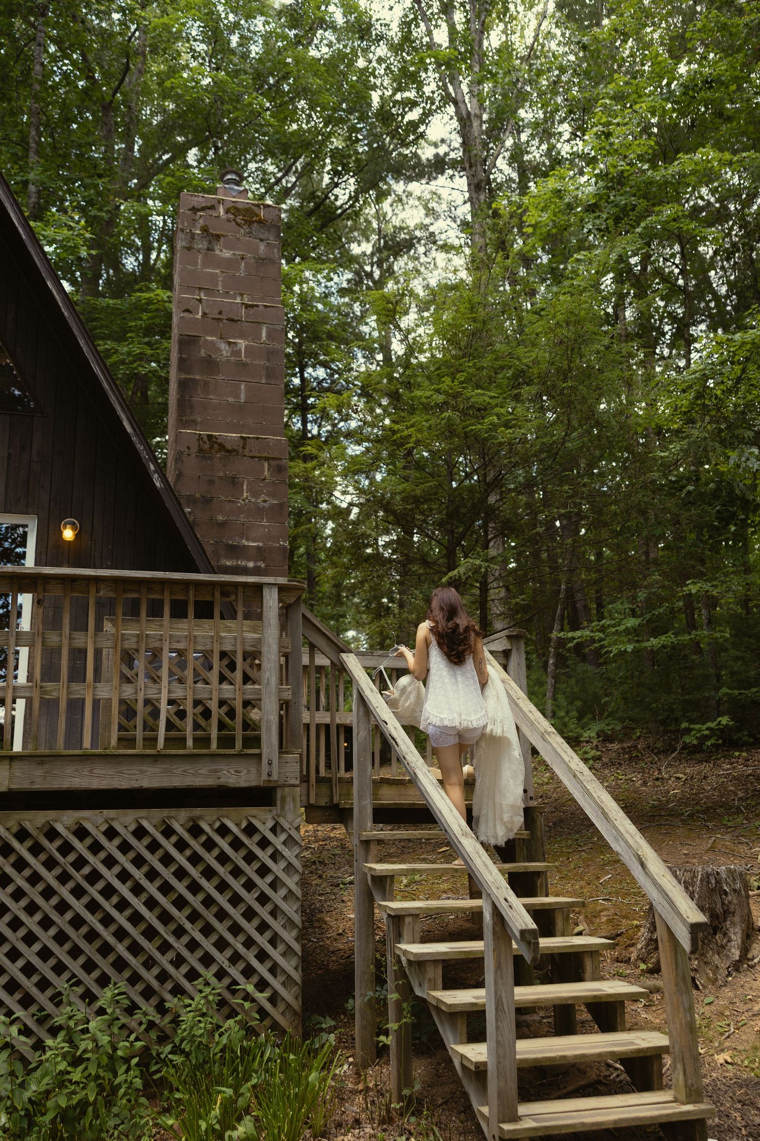 Woman in white dress climbing wooden stairs to a rustic forest cabin with brick chimney.