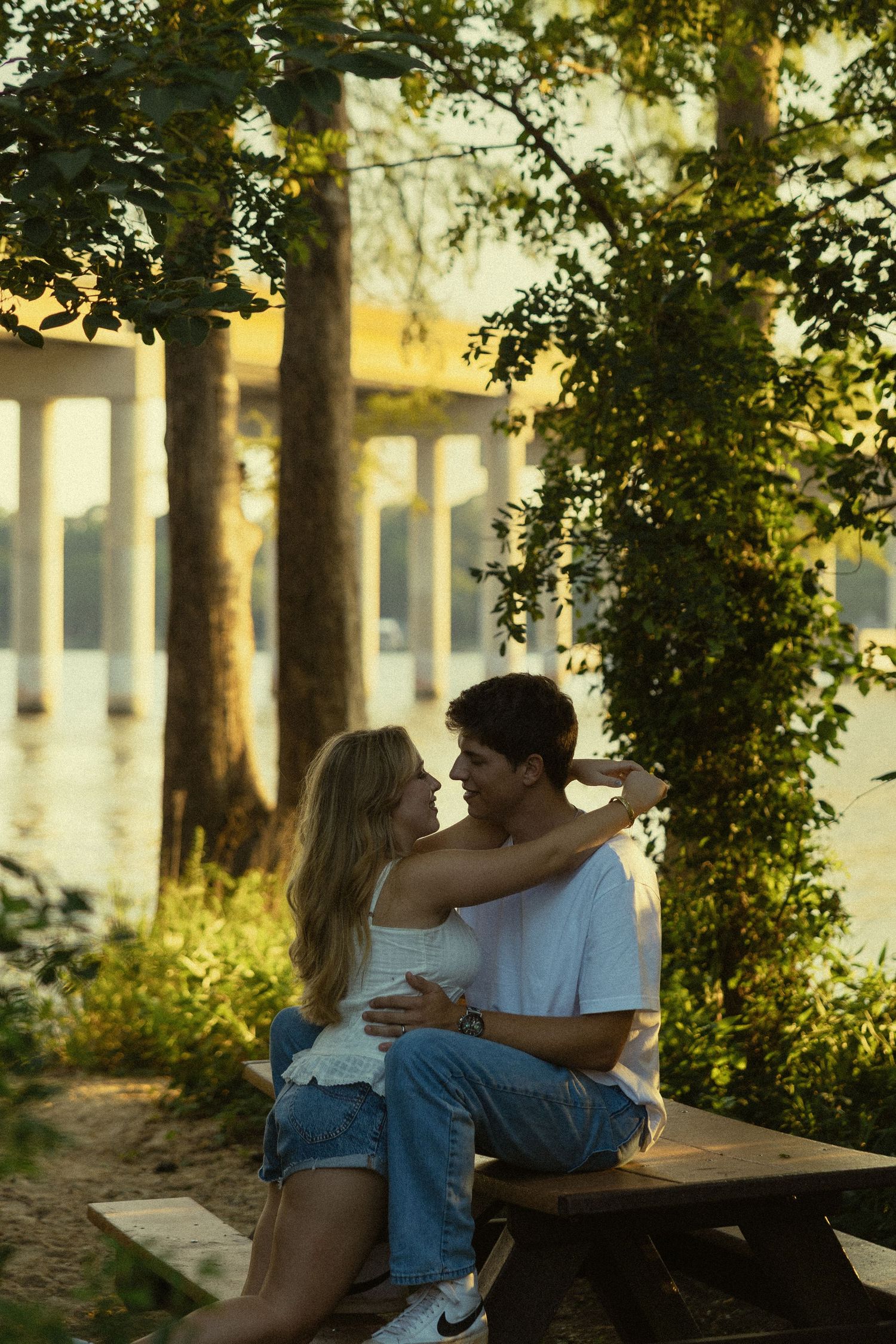 A couple shares a romantic moment sitting together by a sunlit riverside park, surrounded by lush green trees.