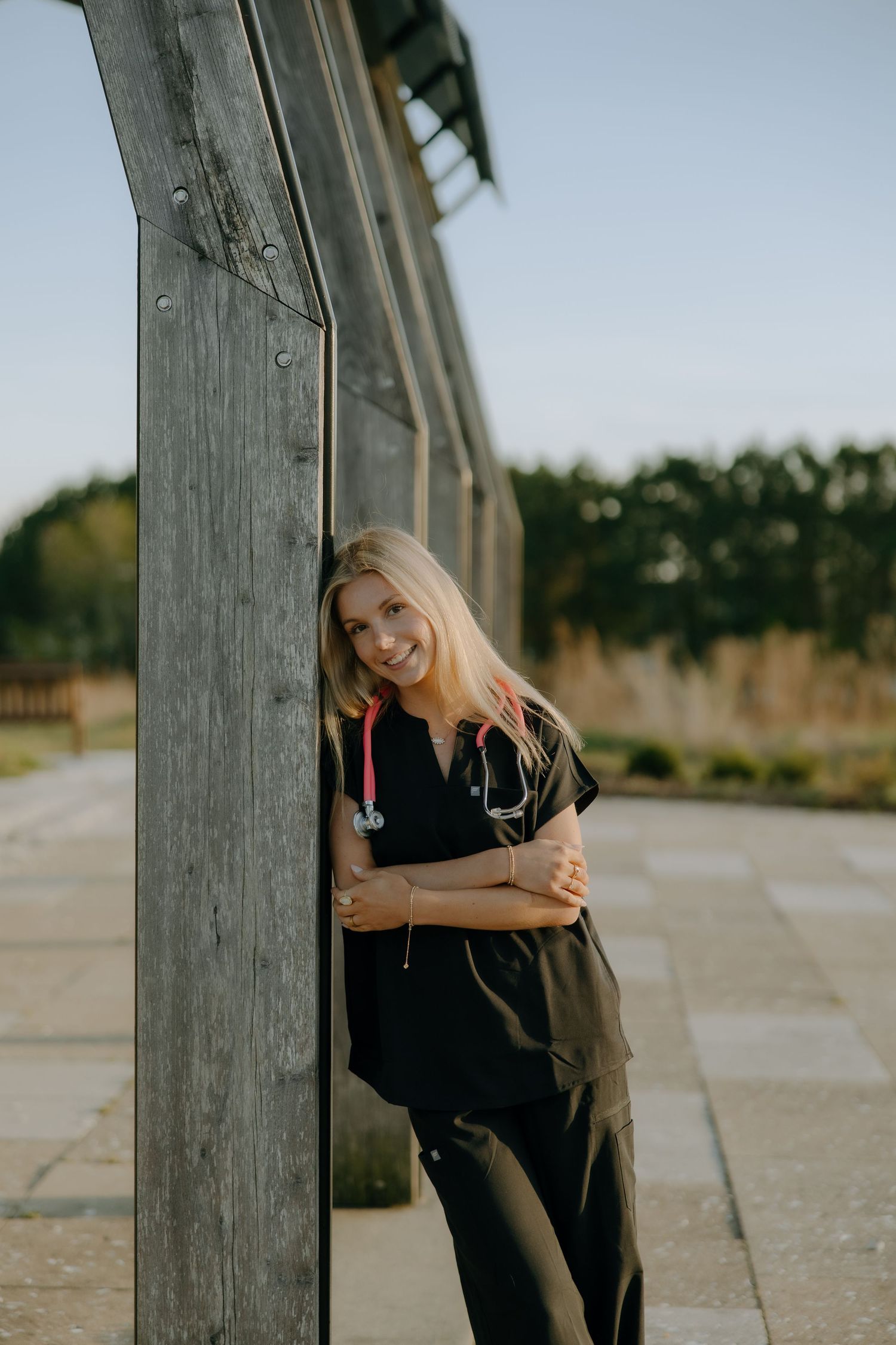Smiling blonde nurse in black scrubs with stethoscope leaning against wooden post outdoors.