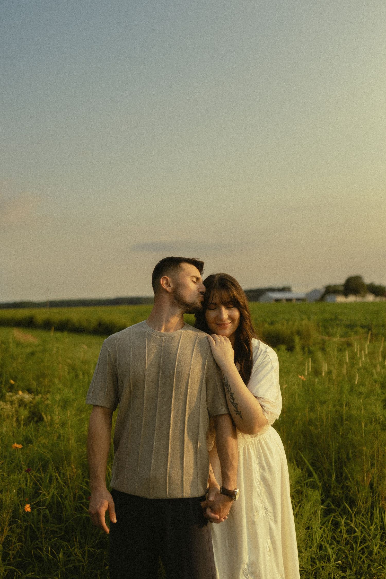 A couple shares a tender moment in a green field at golden hour, the man kissing the woman's head as she smiles softly.