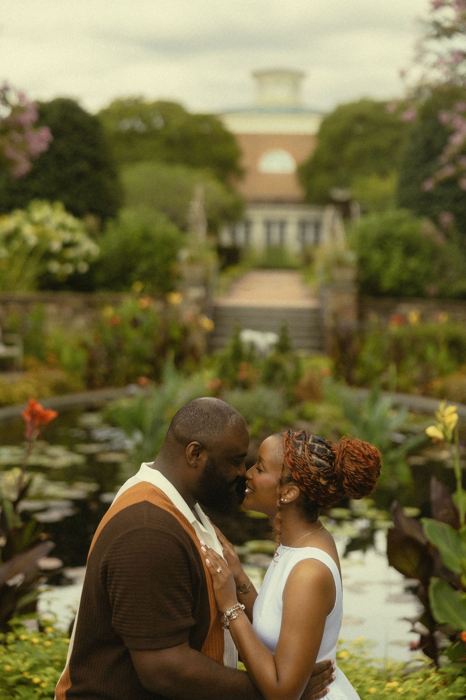 Couple sharing a romantic moment in a lush garden with a pond and elegant building backdrop.