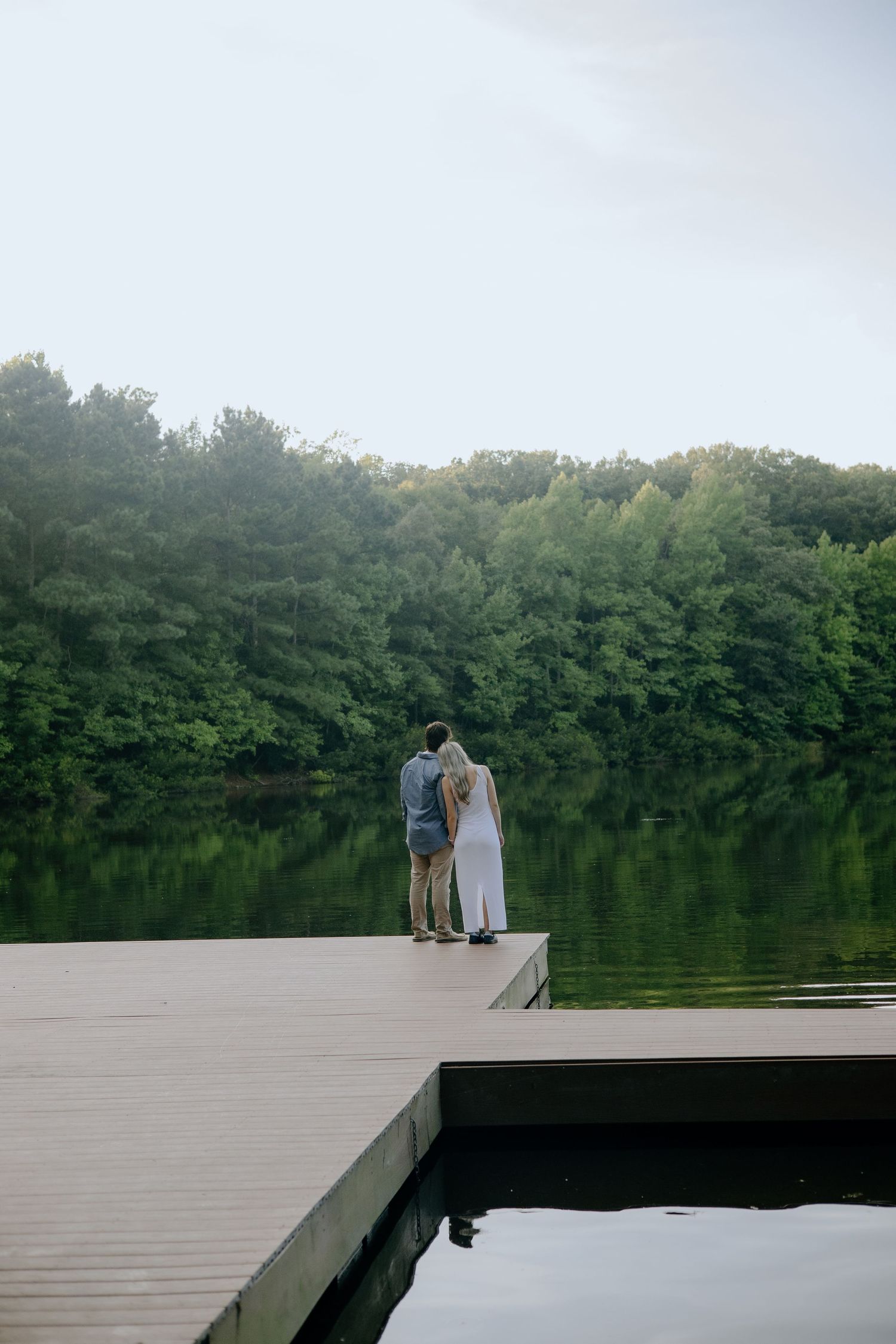 A couple sits together on a wooden dock overlooking a serene lake surrounded by trees.