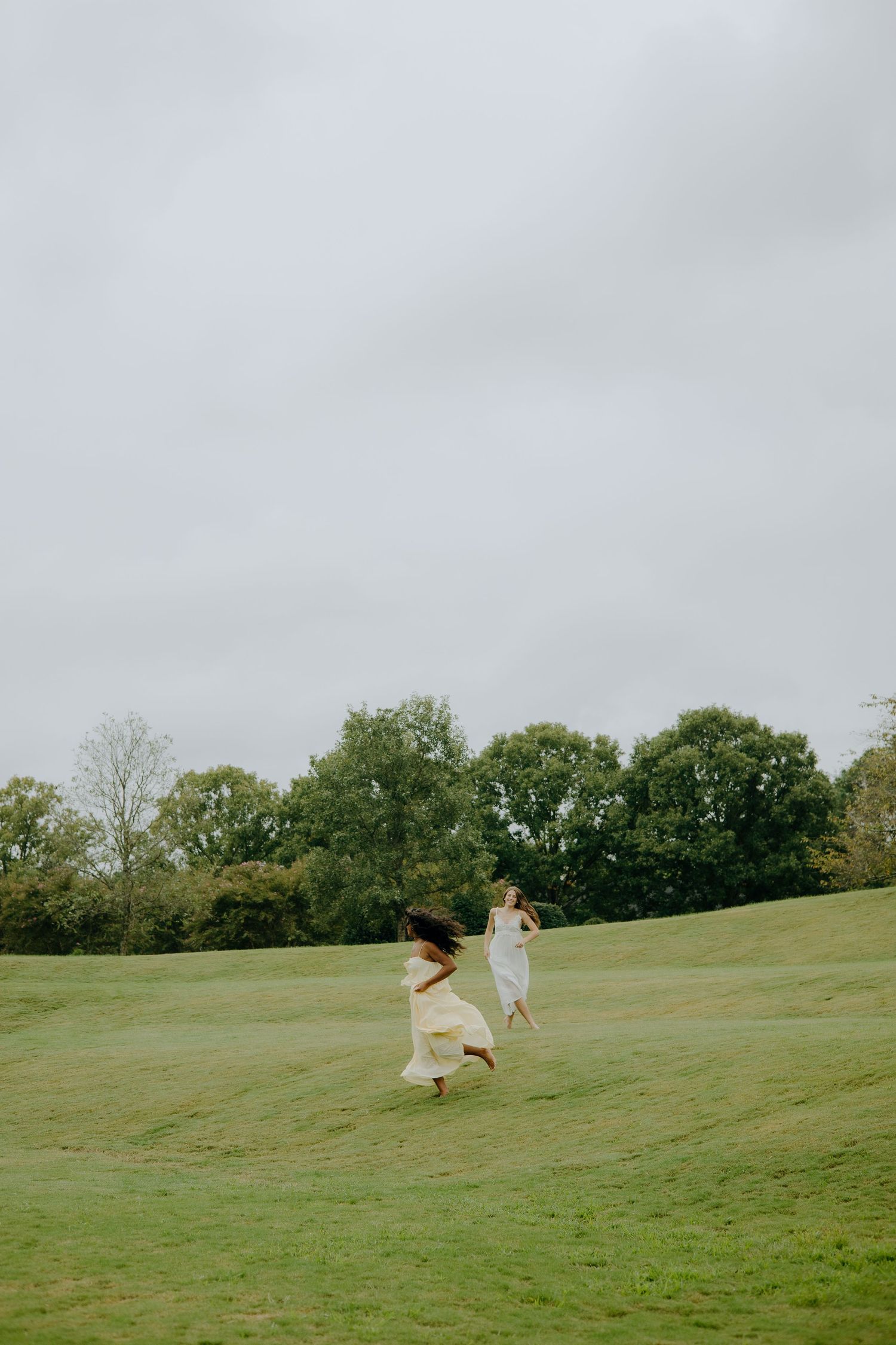 People in flowing dresses run joyfully across a green hillside under cloudy skies.