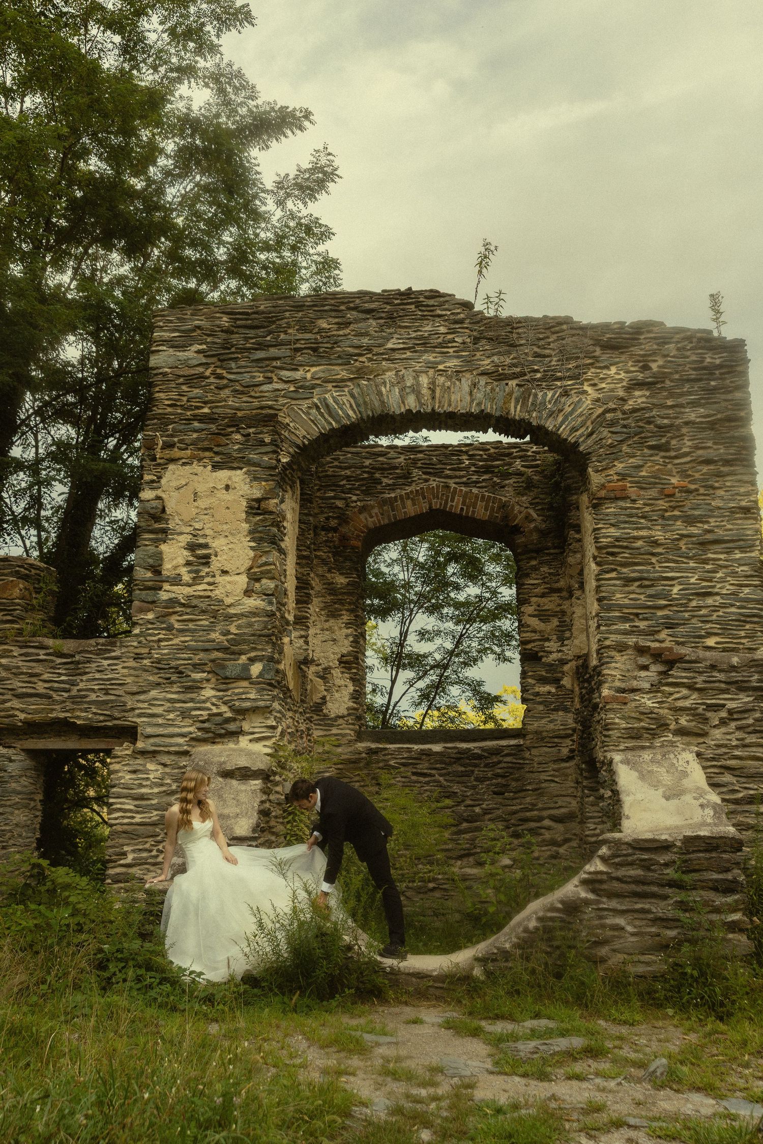 Bride and groom posing romantically among ancient stone ruins with arched window and lush trees.
