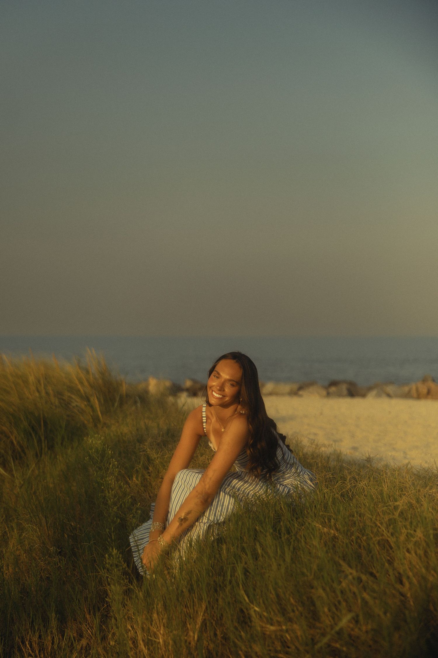 A smiling young woman in a striped dress sits in golden beach grass at sunset with the ocean in the background.