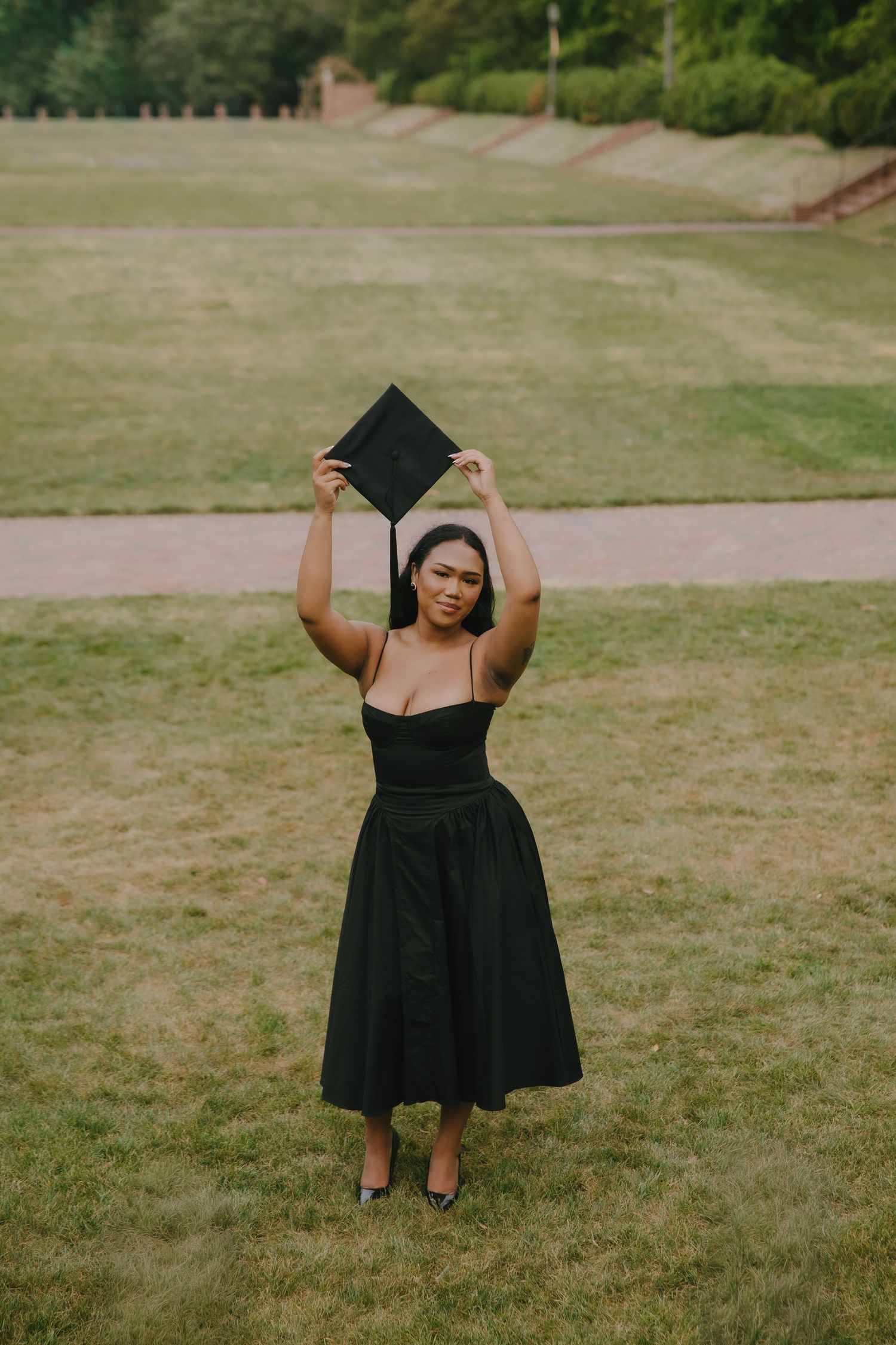 Young woman in black dress holding graduation cap overhead on green campus lawn.