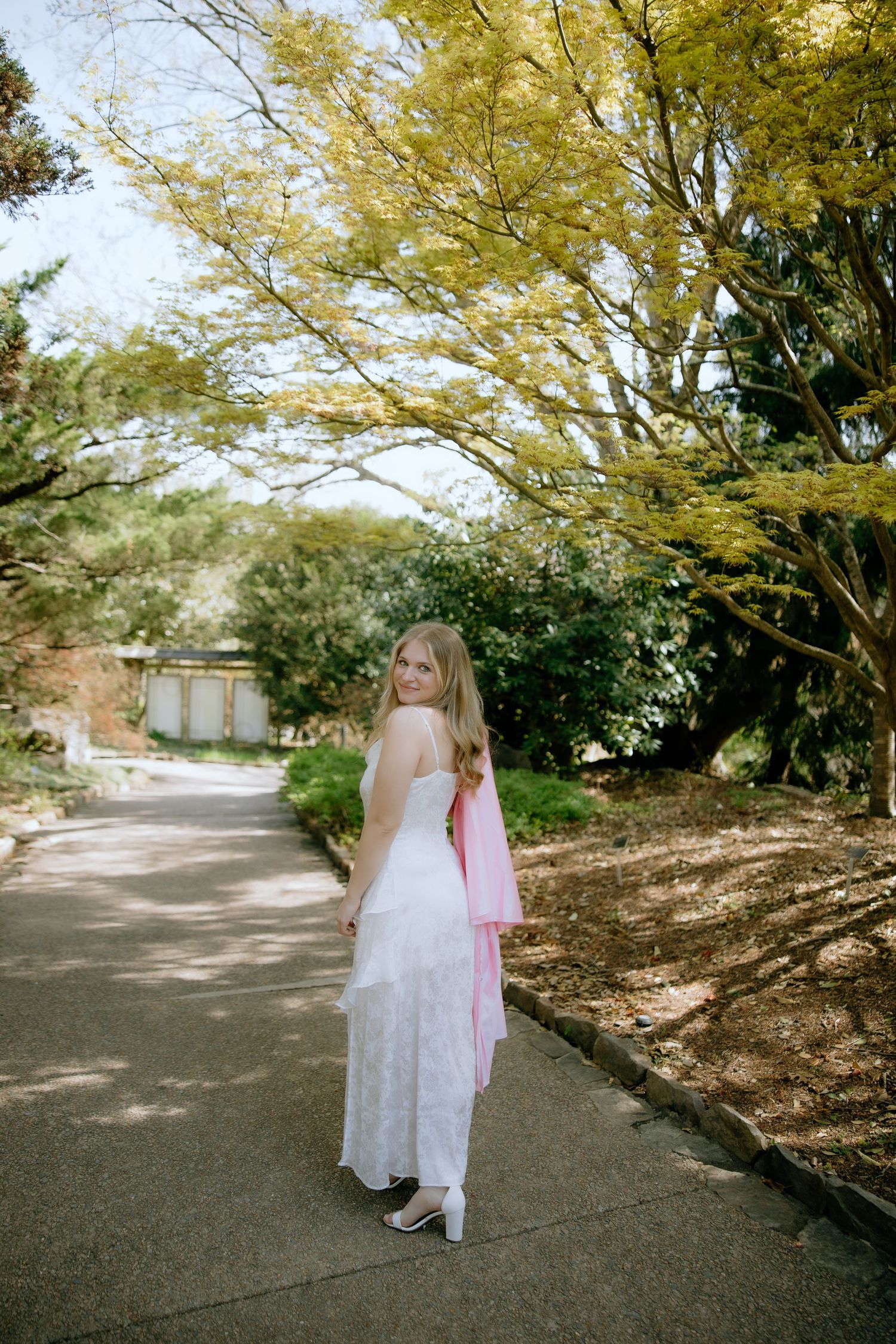 Young woman in white lace dress and pink shawl poses on garden path surrounded by lush green trees.