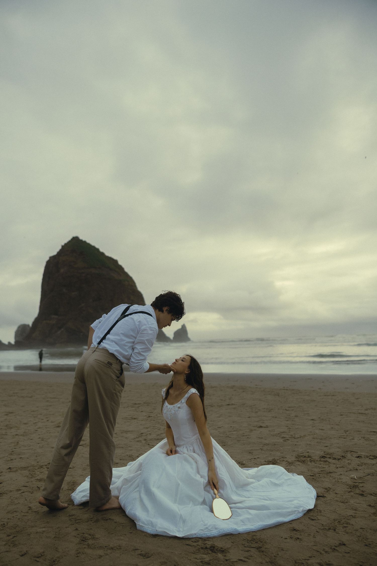 Groom tenderly touches bride's face as she sits on sandy beach with sea stack in background.