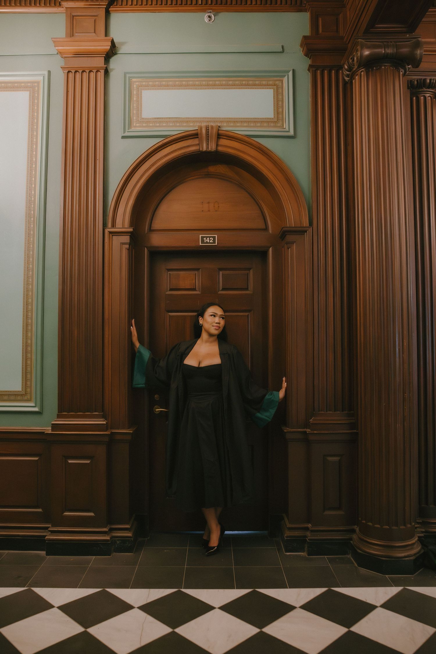 Woman in black graduation gown posing in front of ornate wooden arched door with green walls and checkered floor.