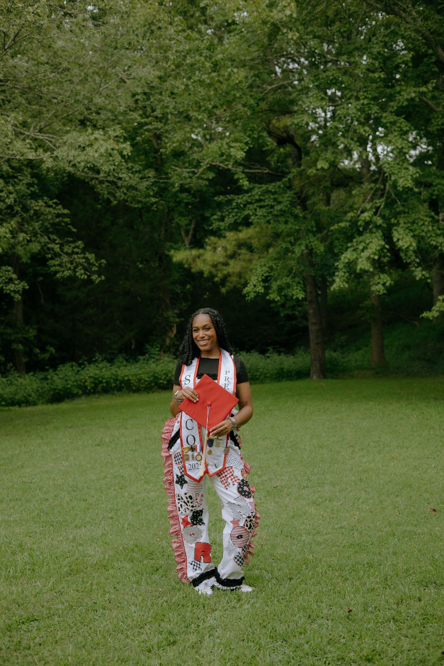 A graduate poses outdoors in white patterned outfit holding red cap with colorful stole in lush green park setting.