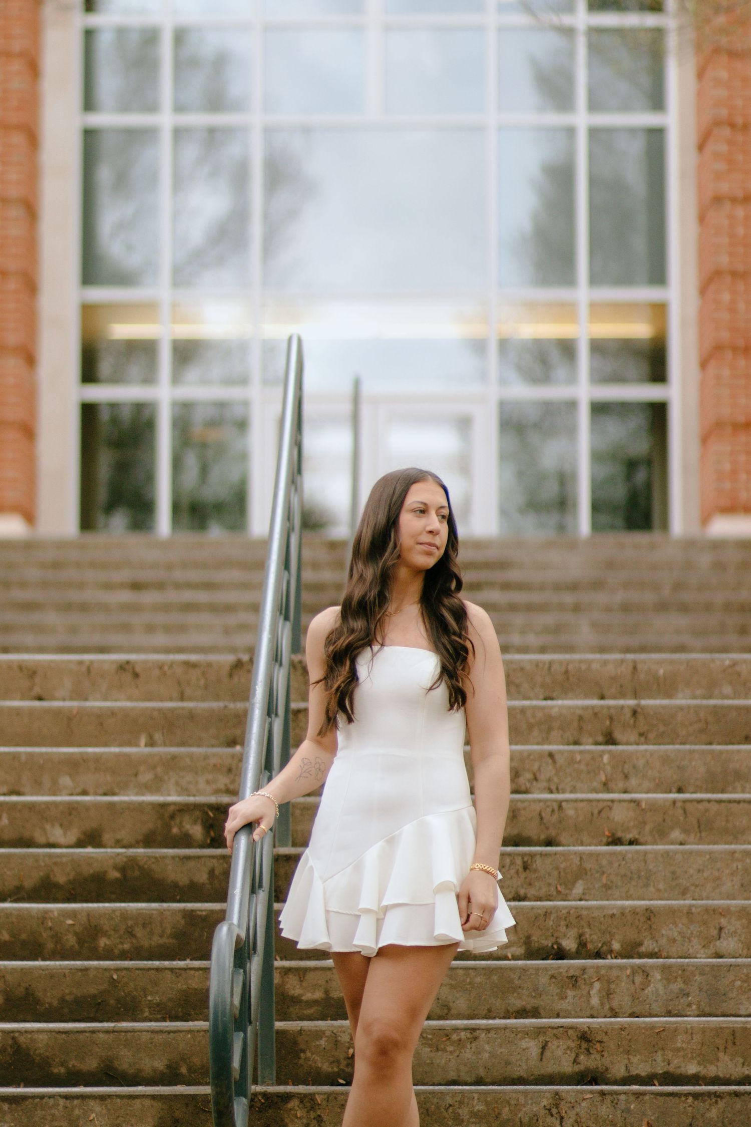 Graduate in white dress stands on steps in front of brick building with large windows.