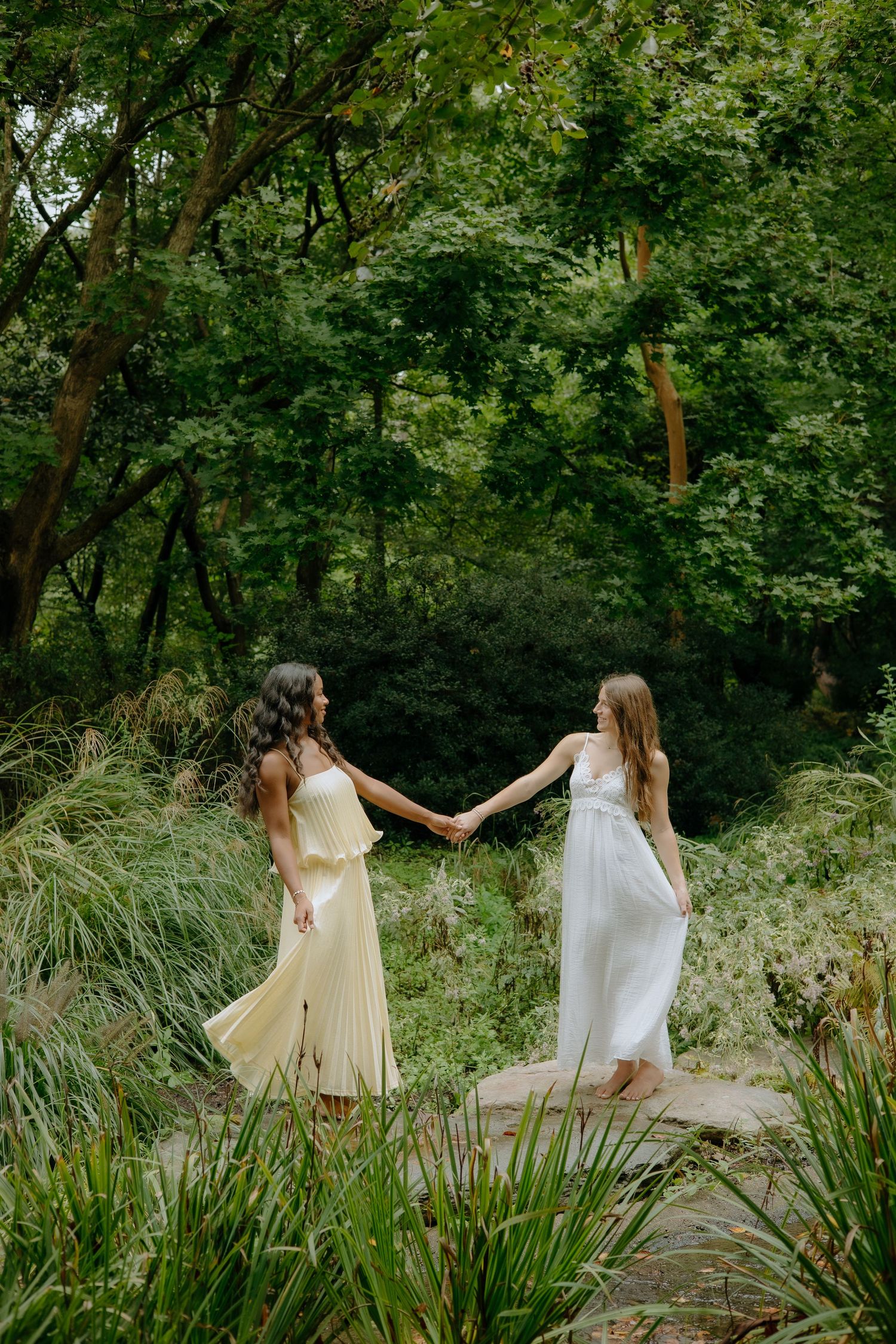 Friends in flowing white dresses dance together in a lush garden setting.