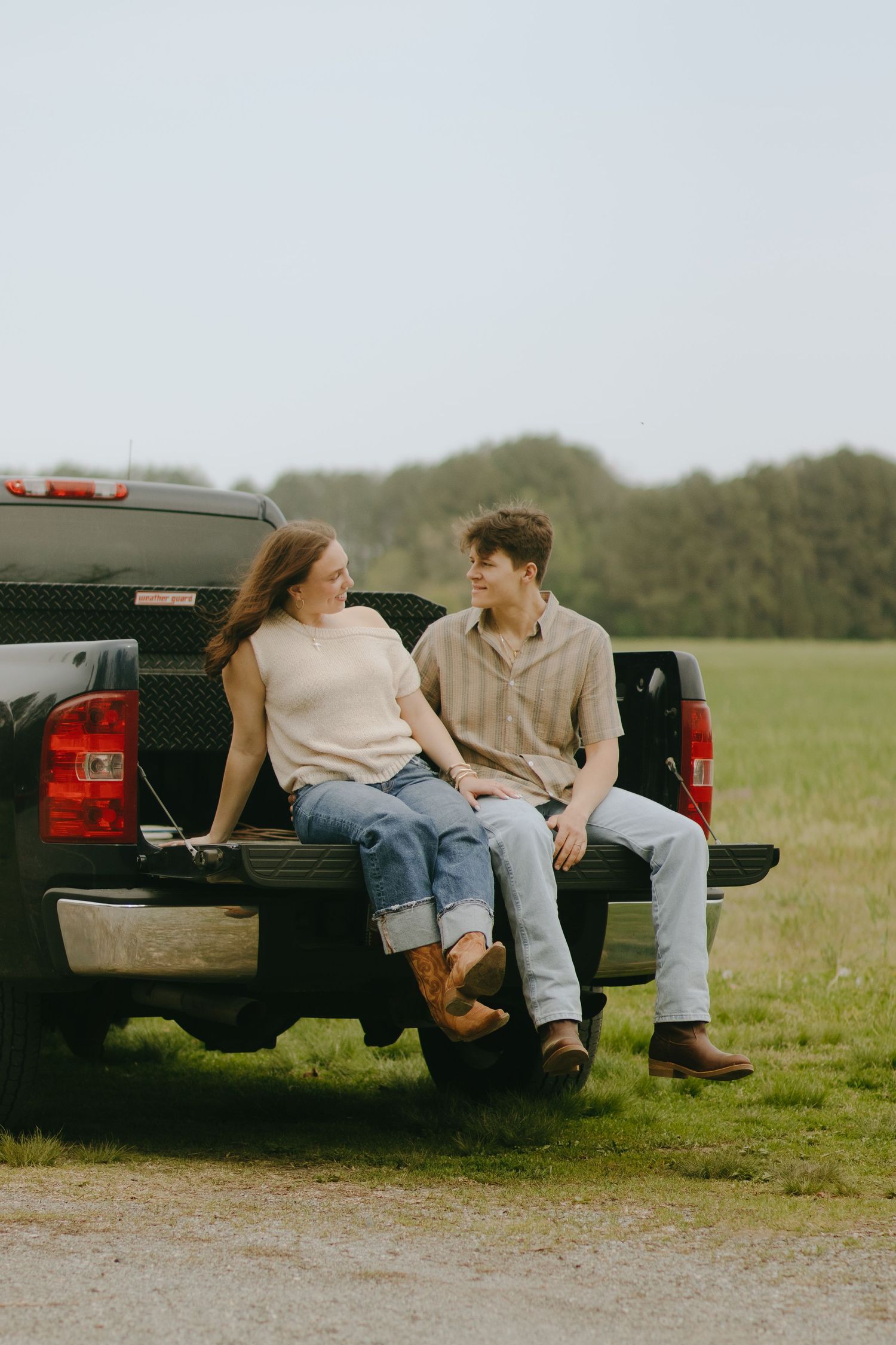 A couple sits together on the tailgate of a black pickup truck in a rural setting.