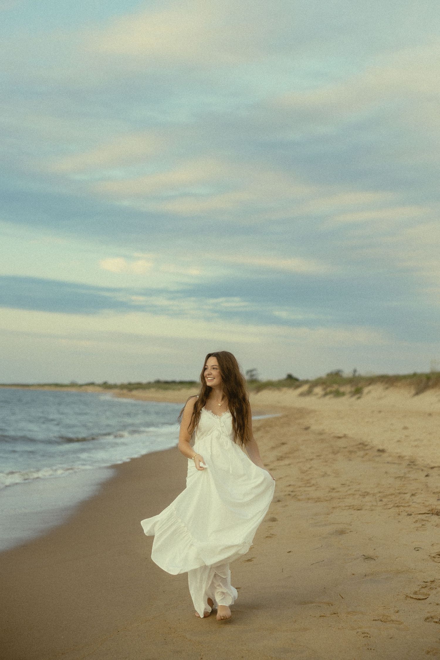 A smiling woman in a flowing white dress walks along a sandy beach at dusk with gentle waves and cloudy sky.