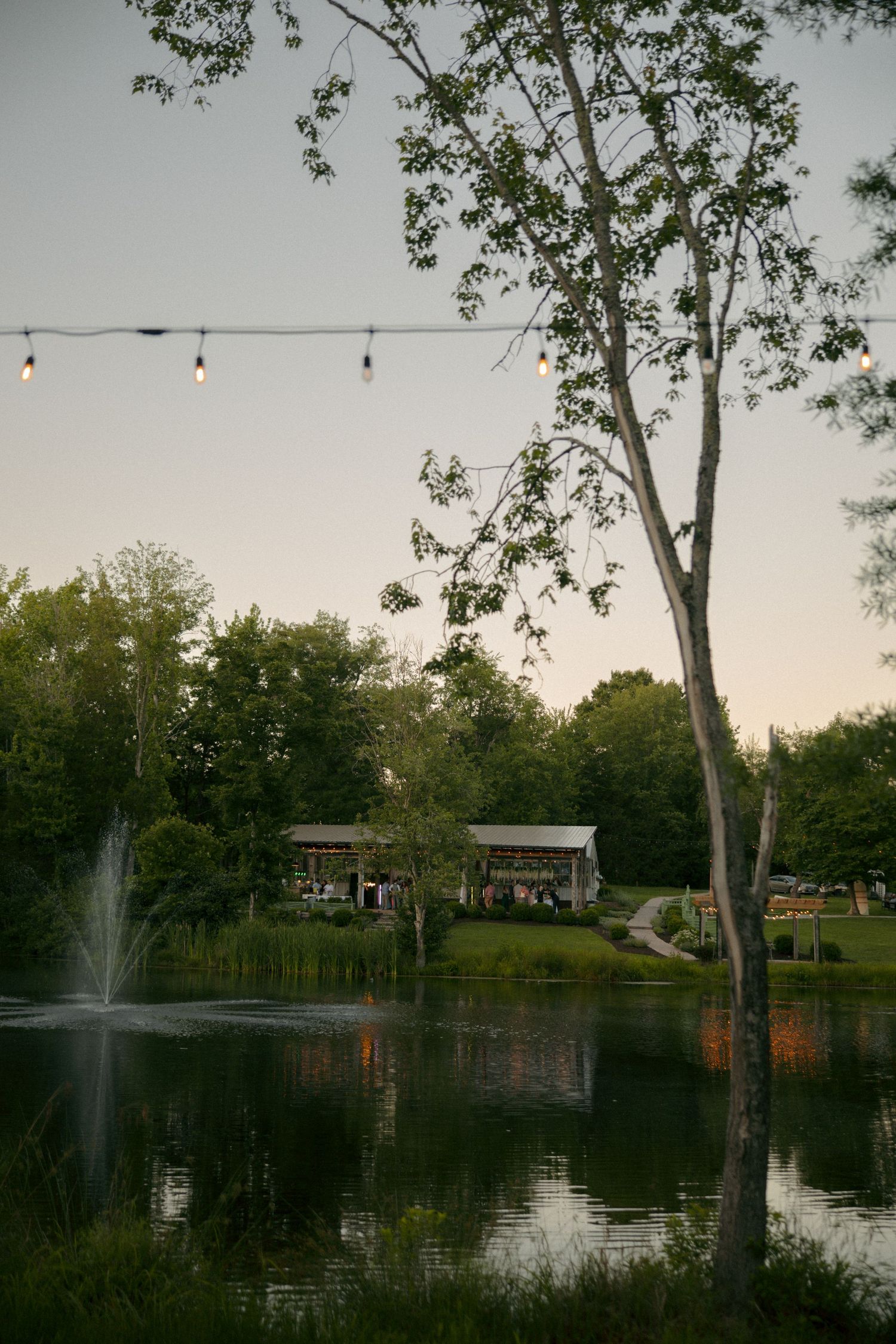Serene pond with fountain, string lights, and rustic pavilion surrounded by lush green trees at dusk.