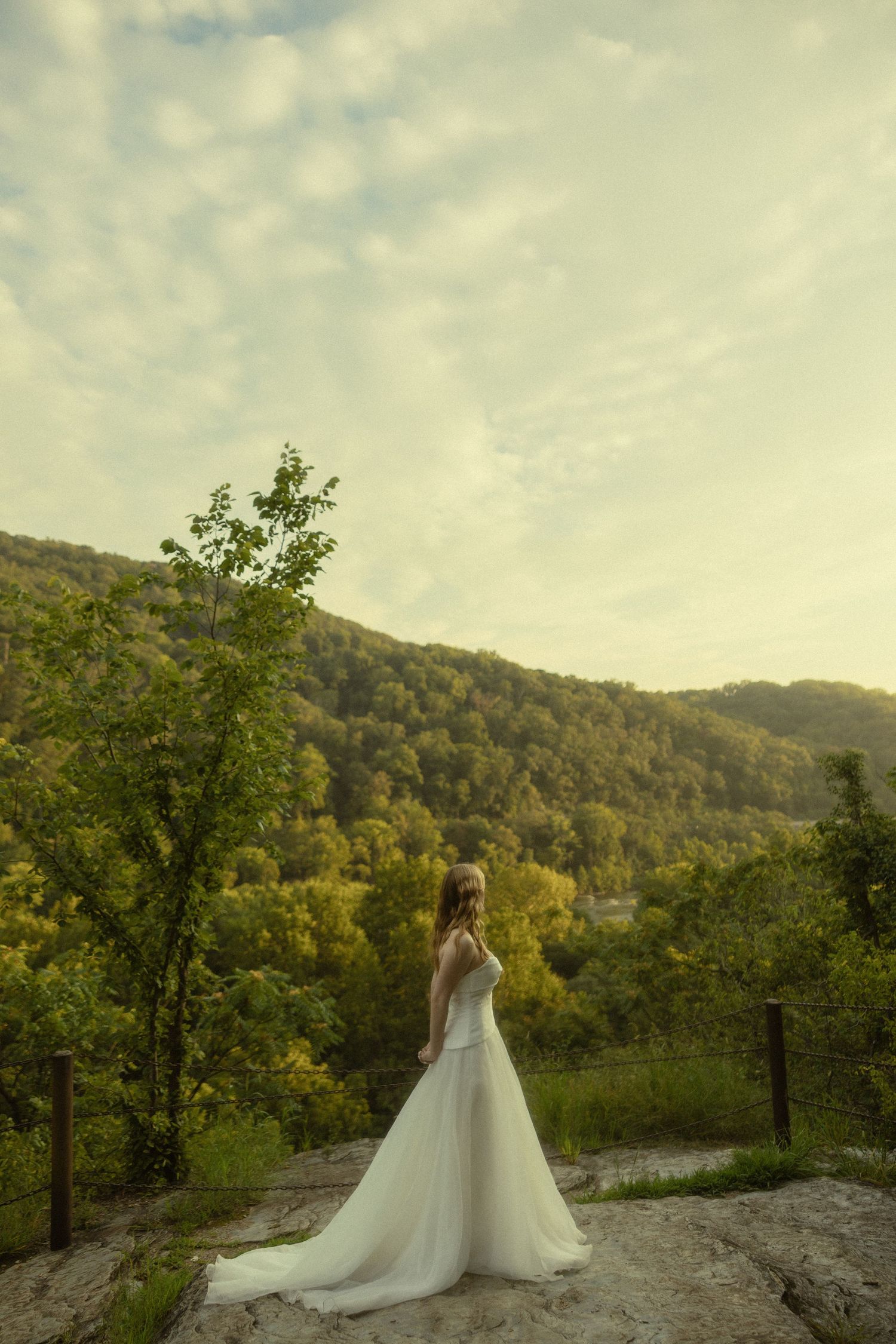 Bride in white gown stands on rocky overlook with lush green forested hills and golden sky behind her.