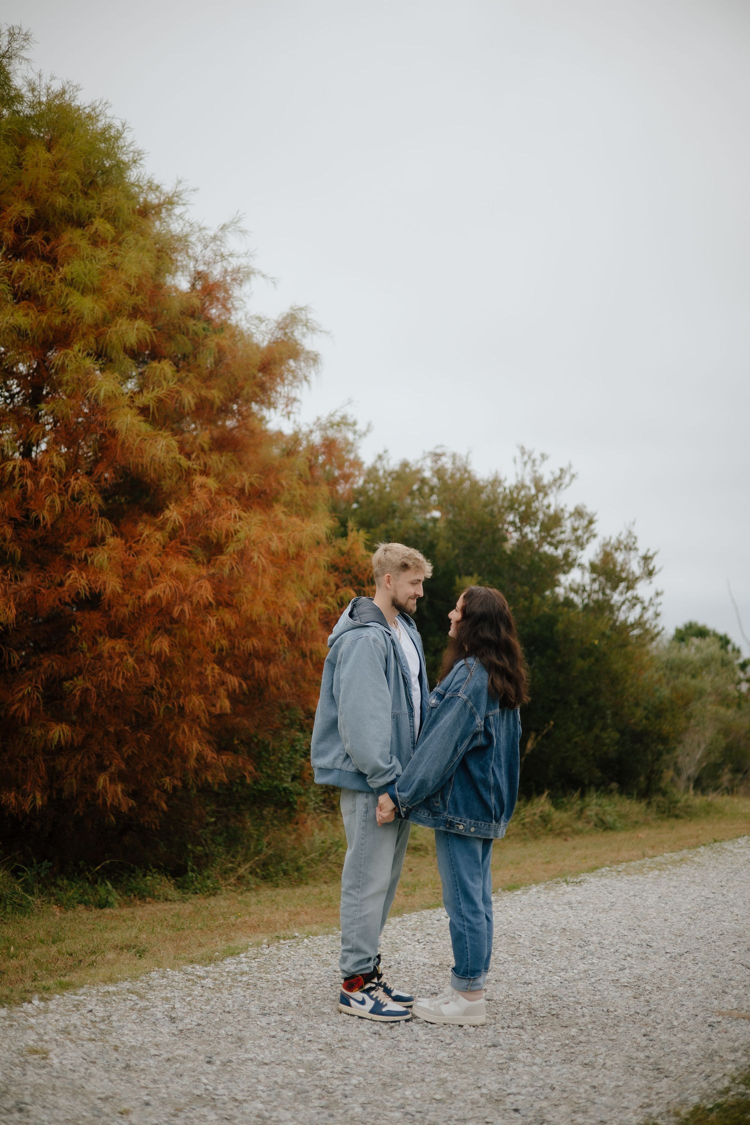 Two people in casual denim outfits standing together on a gravel path in autumn.