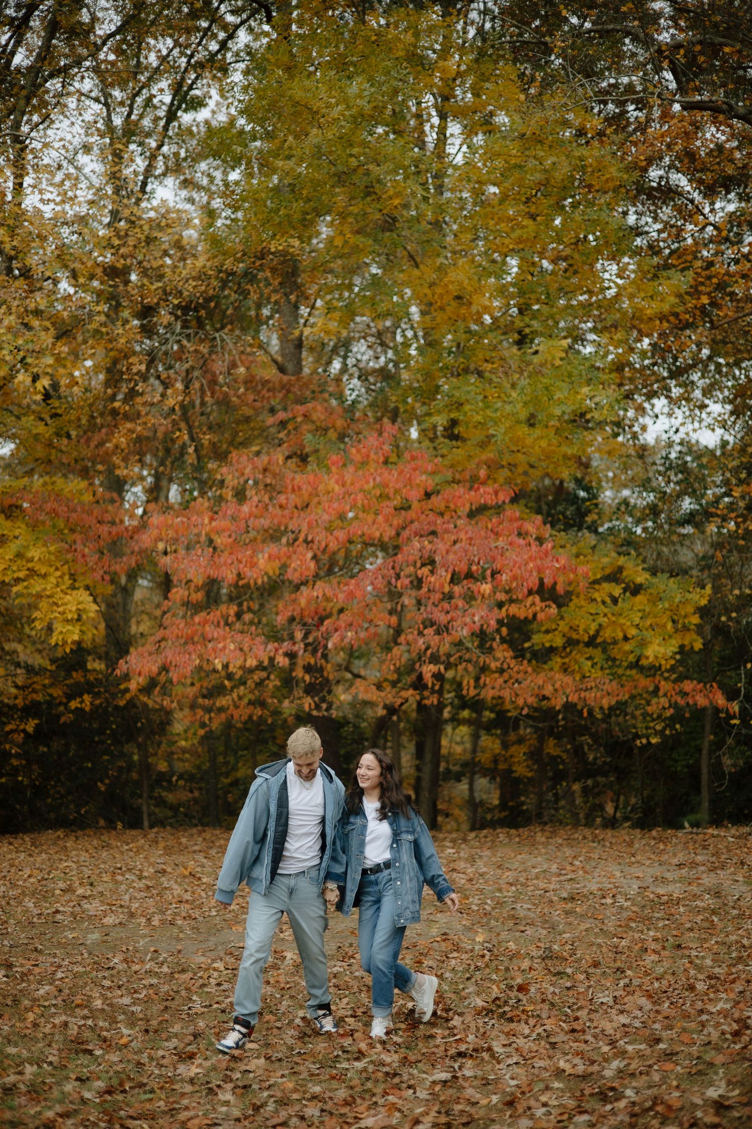 A couple walks together through fallen leaves beneath vibrant autumn trees with red and gold foliage.