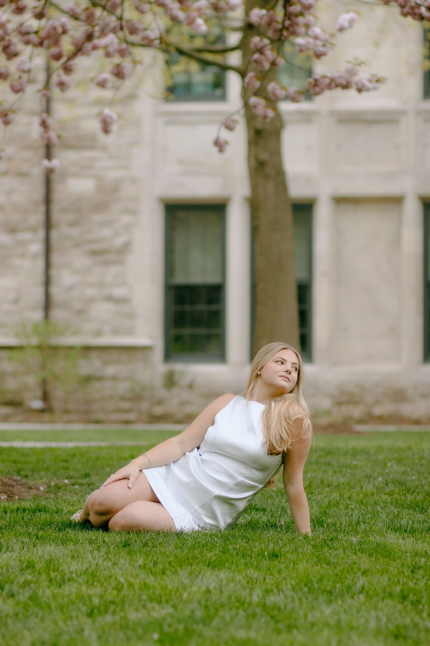 Reclining pose on green grass with blooming cherry blossoms and stone architecture in background.