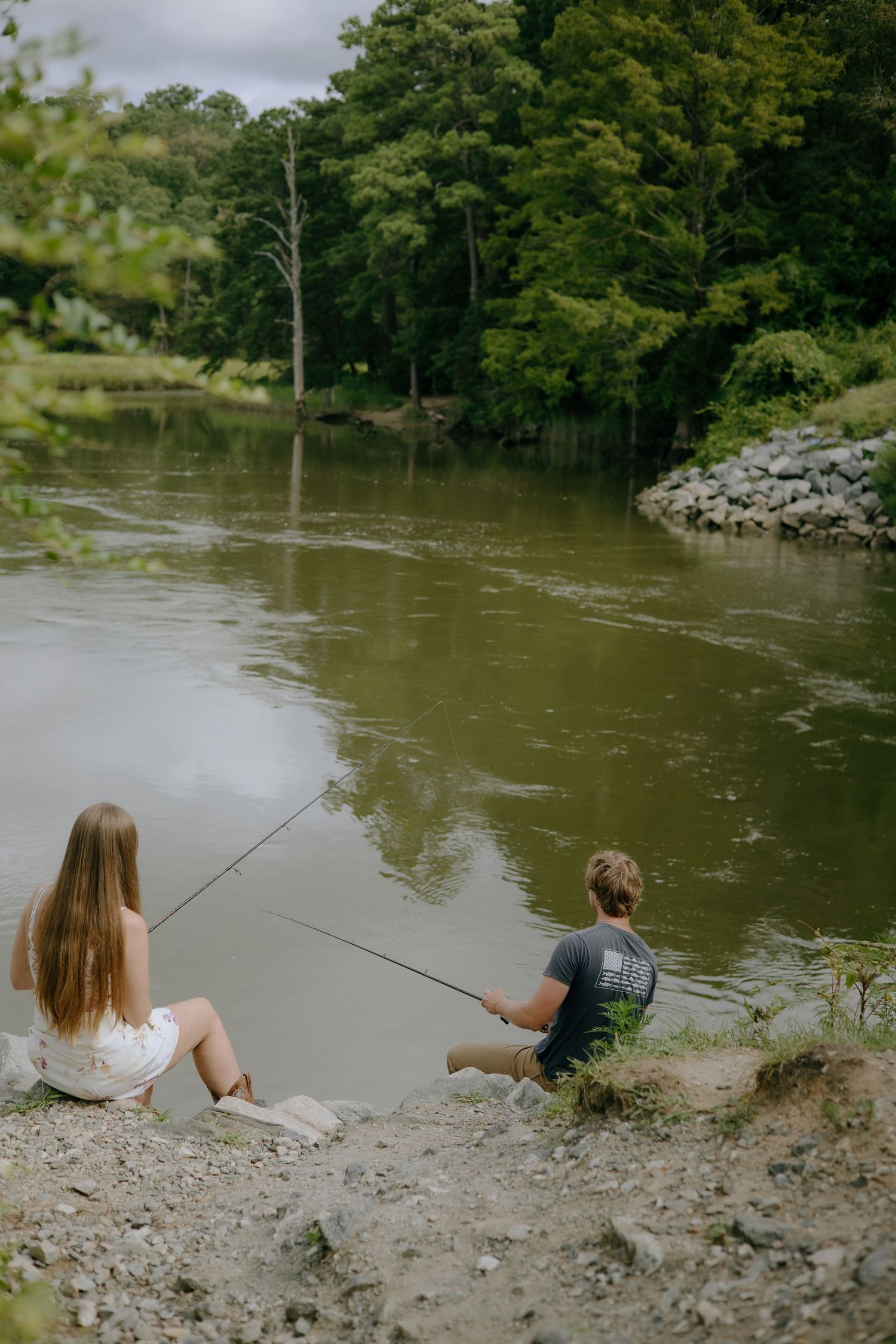 A couple sits together on a riverbank fishing on a cloudy day near trees and rocks.
