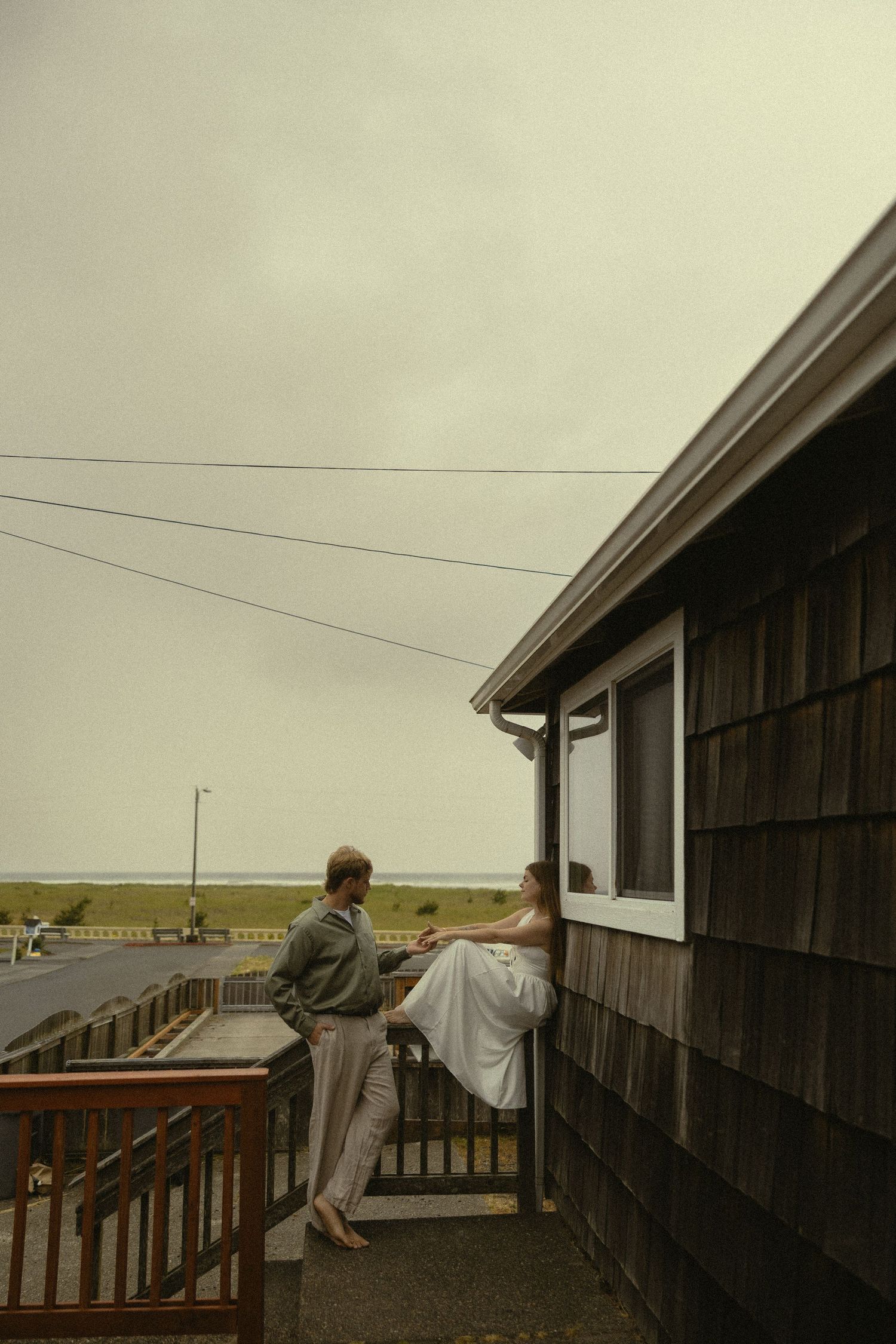 Couple on a coastal cottage deck, woman in white dress sitting by window with ocean view on overcast day.
