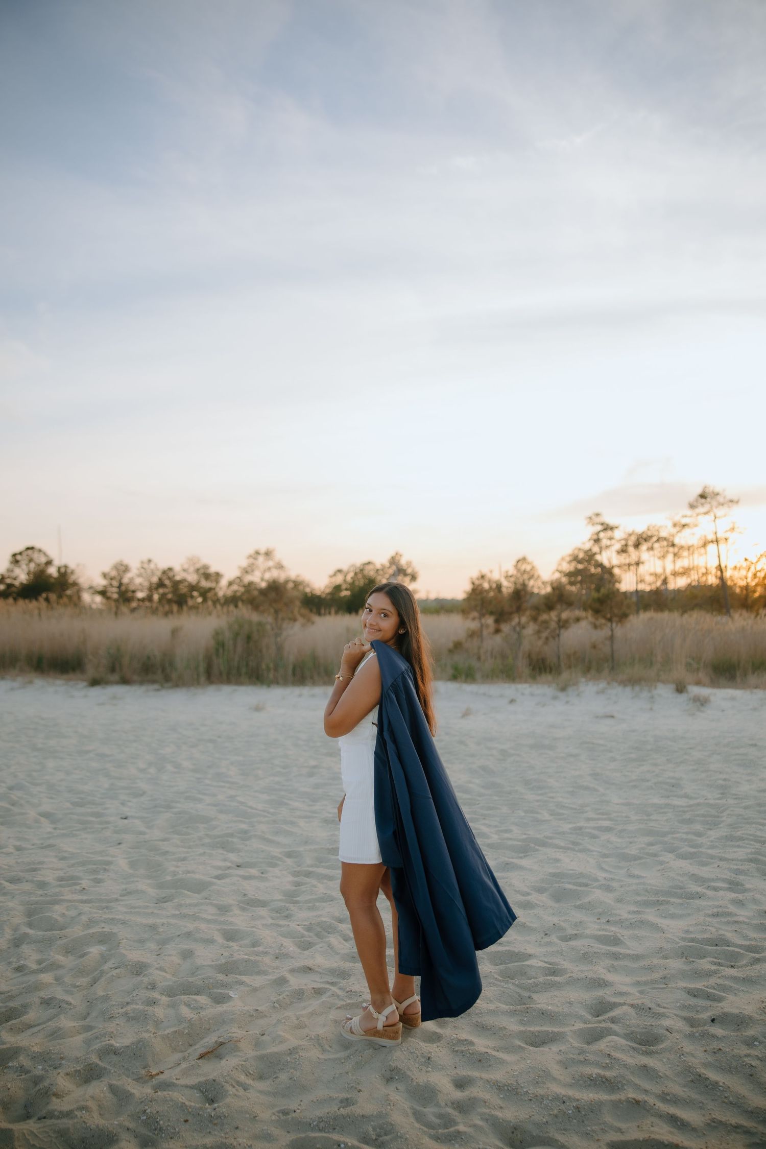 Woman in white dress holding navy blue scarf on sandy beach at sunset with trees in background.