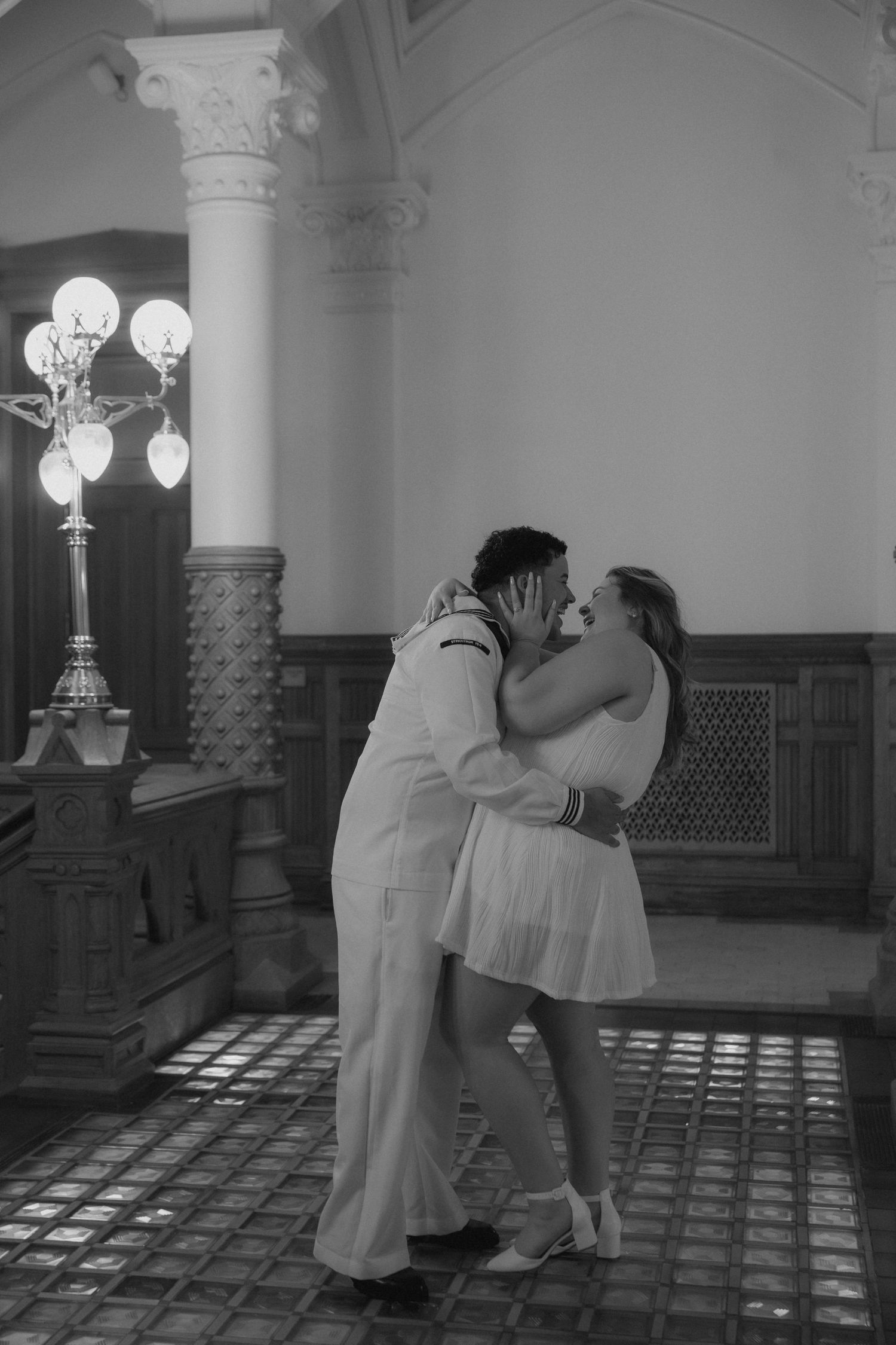 Black and white photo of a couple sharing a romantic kiss in a grand architectural interior with ornate details.