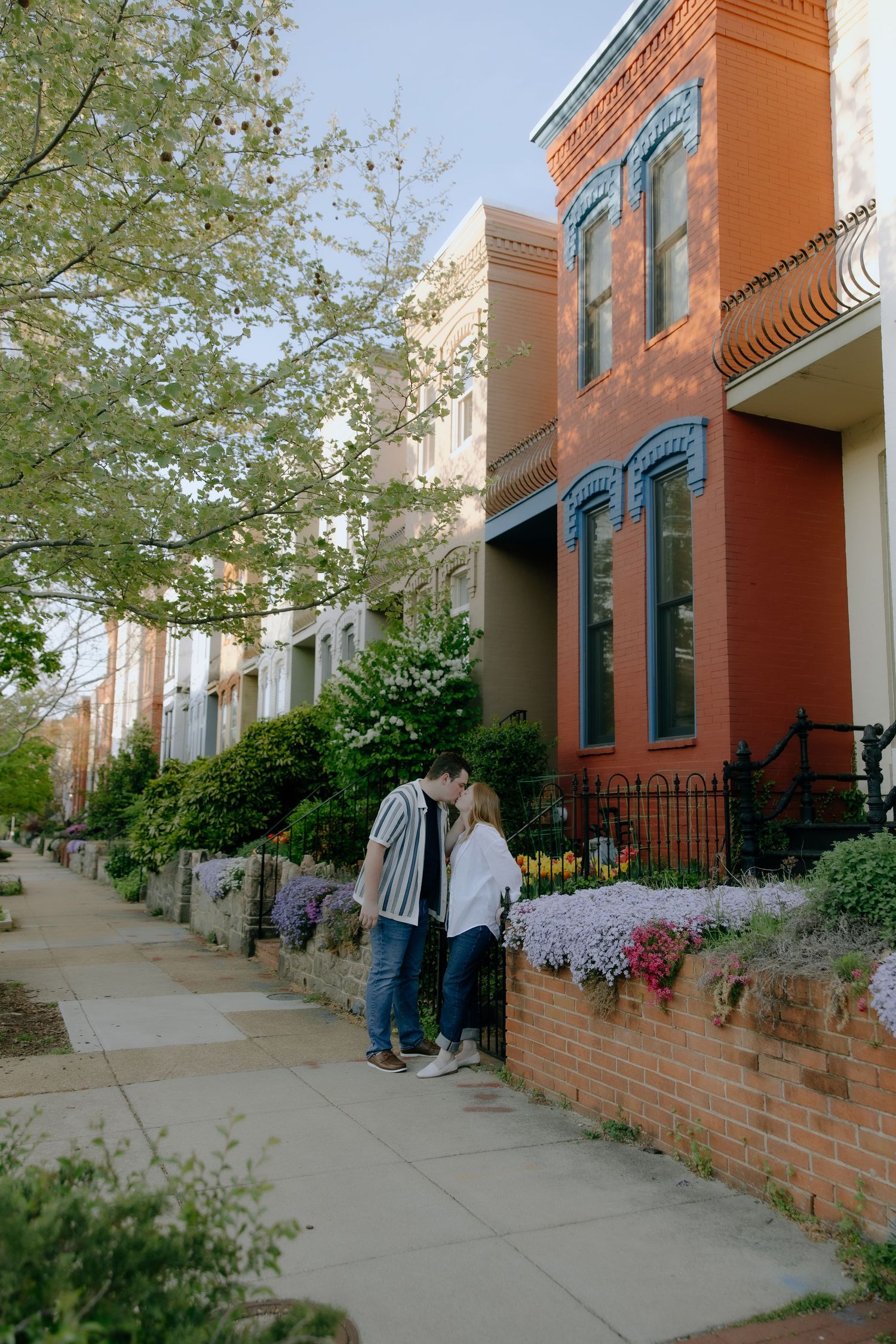 Couple kissing on a charming urban sidewalk lined with colorful row houses and blooming spring flowers.