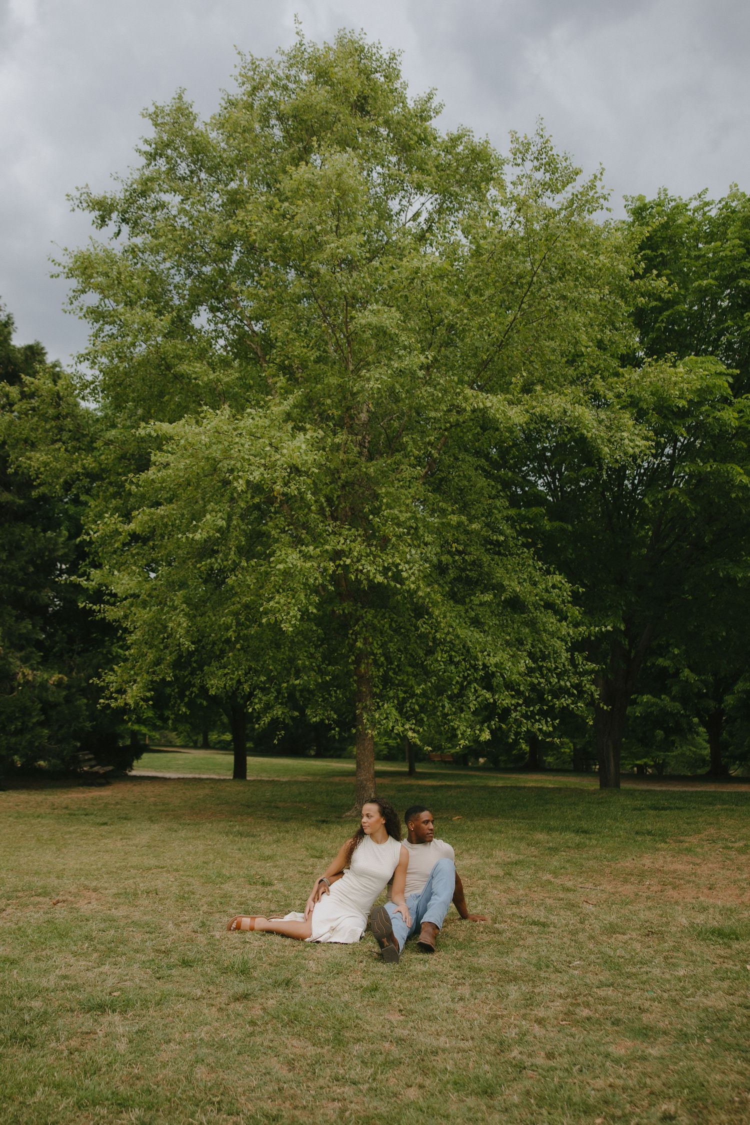An interracial couple sits back-to-back on grass beneath a large green tree in a serene park setting.
